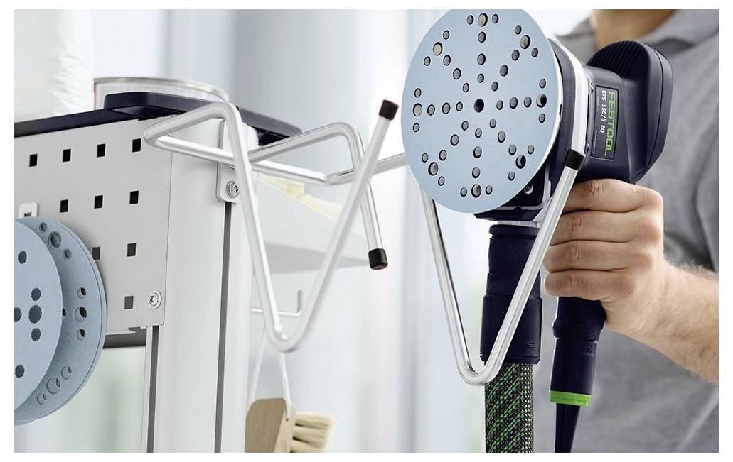 Eine Person hält ein Elektrowerkzeug mit einem runden, perforierten Schleifteller neben einem wandmontierten Werkzeughalter, was auf professionelle Arbeit hindeutet.