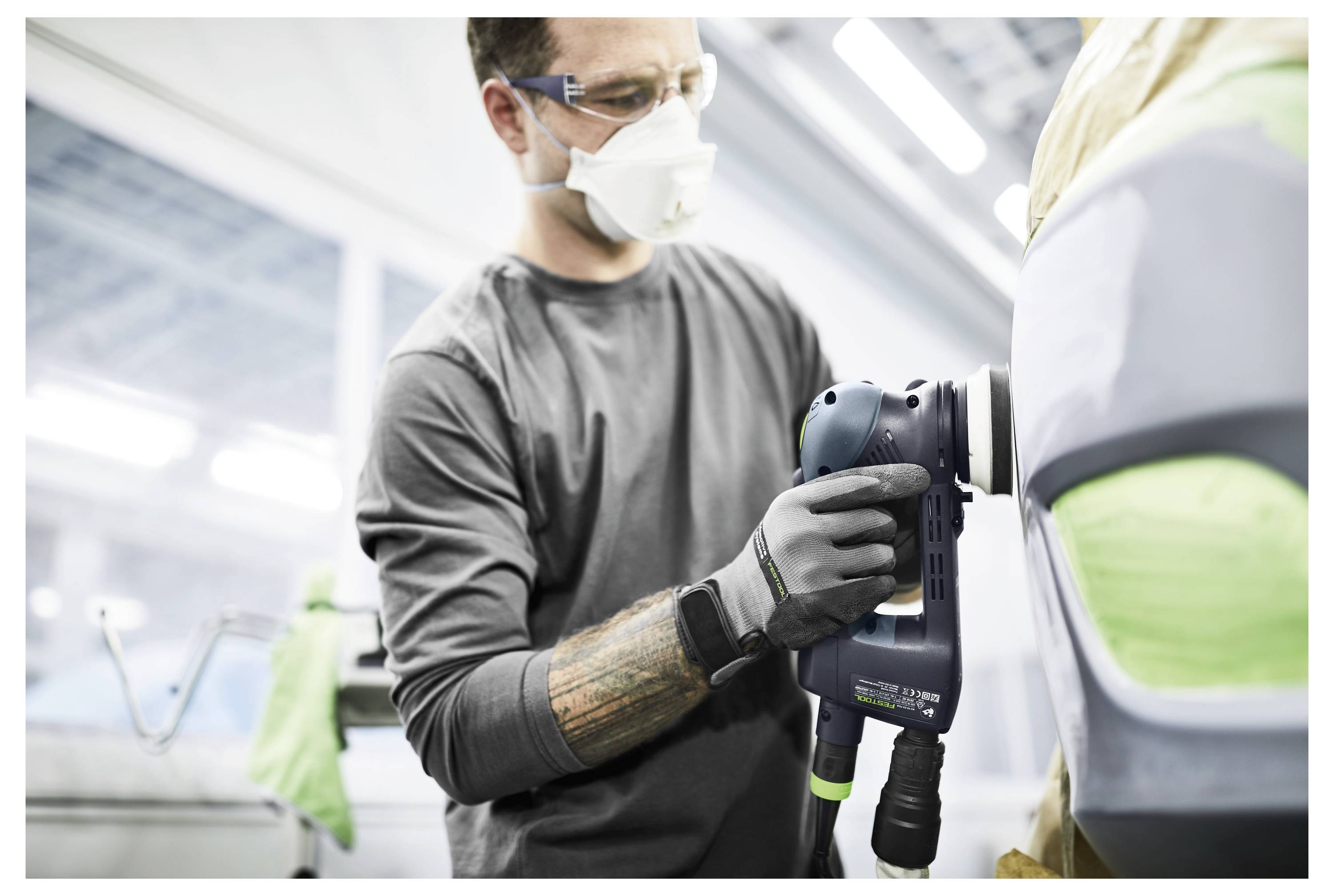 Eine Person in Schutzausrüstung verwendet einen elektrischen Schleiffer auf einer Autoberfläche in einer Werkstatt.