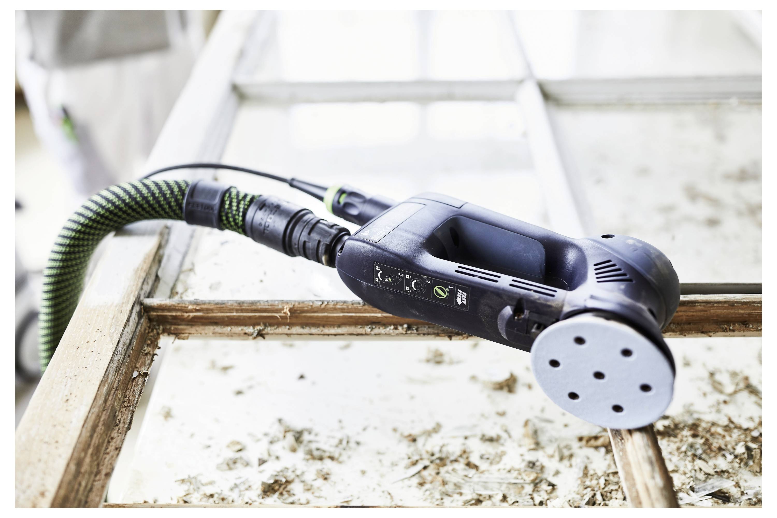 Ein elektrischer Schleiffer liegt auf einem Fensterrahmen und glättet altes Holz. Staub und Verschmutzungen sind sichtbar und deuten auf ein laufendes Restaurierungsprojekt hin.