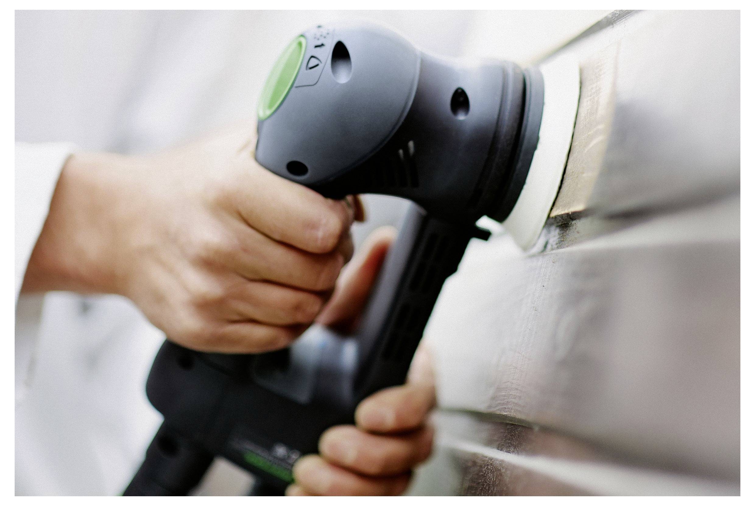 Eine Person benutzt einen elektrischen Schleifer auf einer glatten Oberfläche und konzentriert sich auf gleichmäßiges Schleifen. Das Werkzeug wird mit beiden Händen fest gehalten, um Präzision zu gewährleisten.