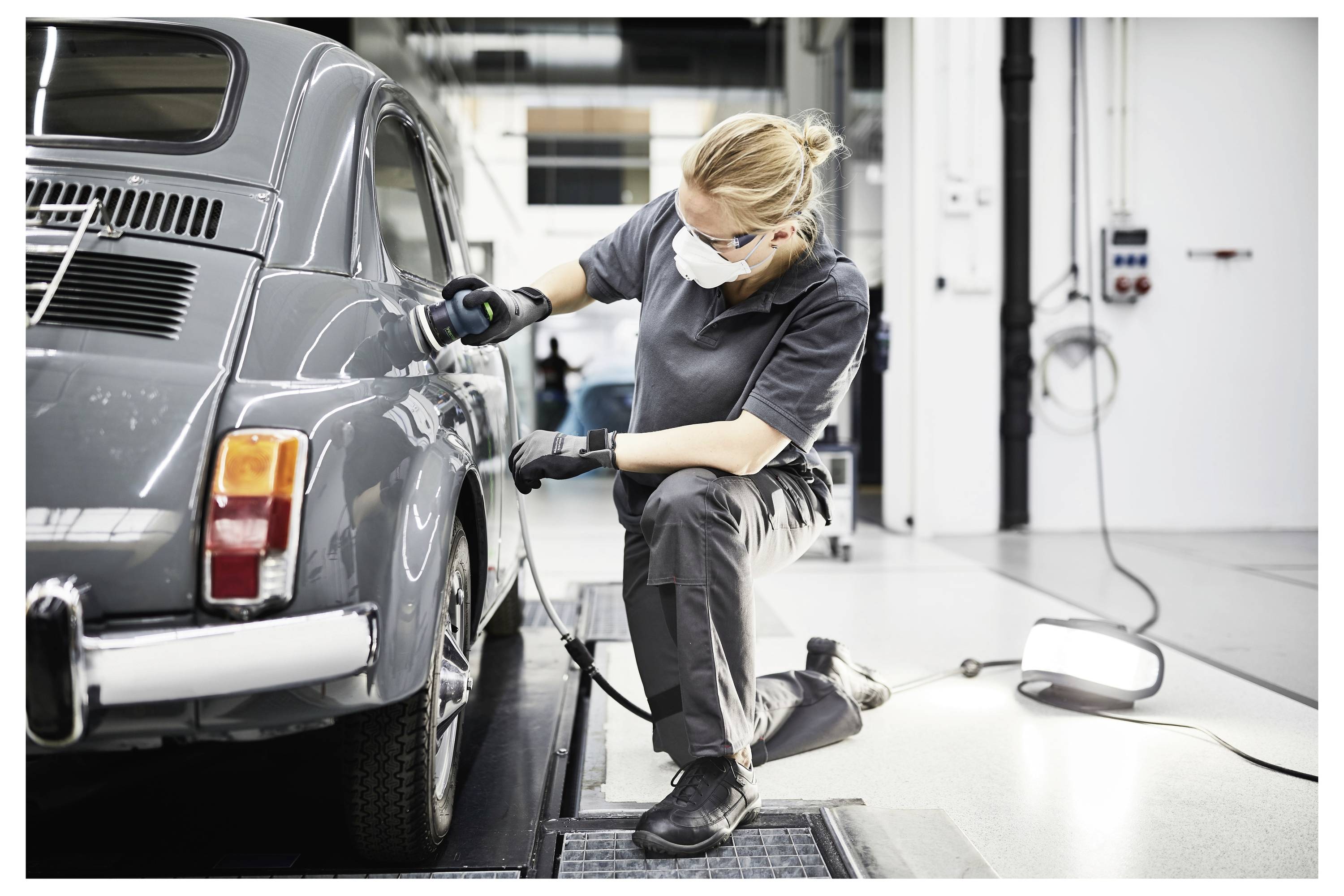 Eine Person, die eine Maske und Handschuhe trägt, poliert ein Oldtimer-Auto in einer hell beleuchteten Garage und konzentriert sich dabei auf das hintere Seitenpaneel.