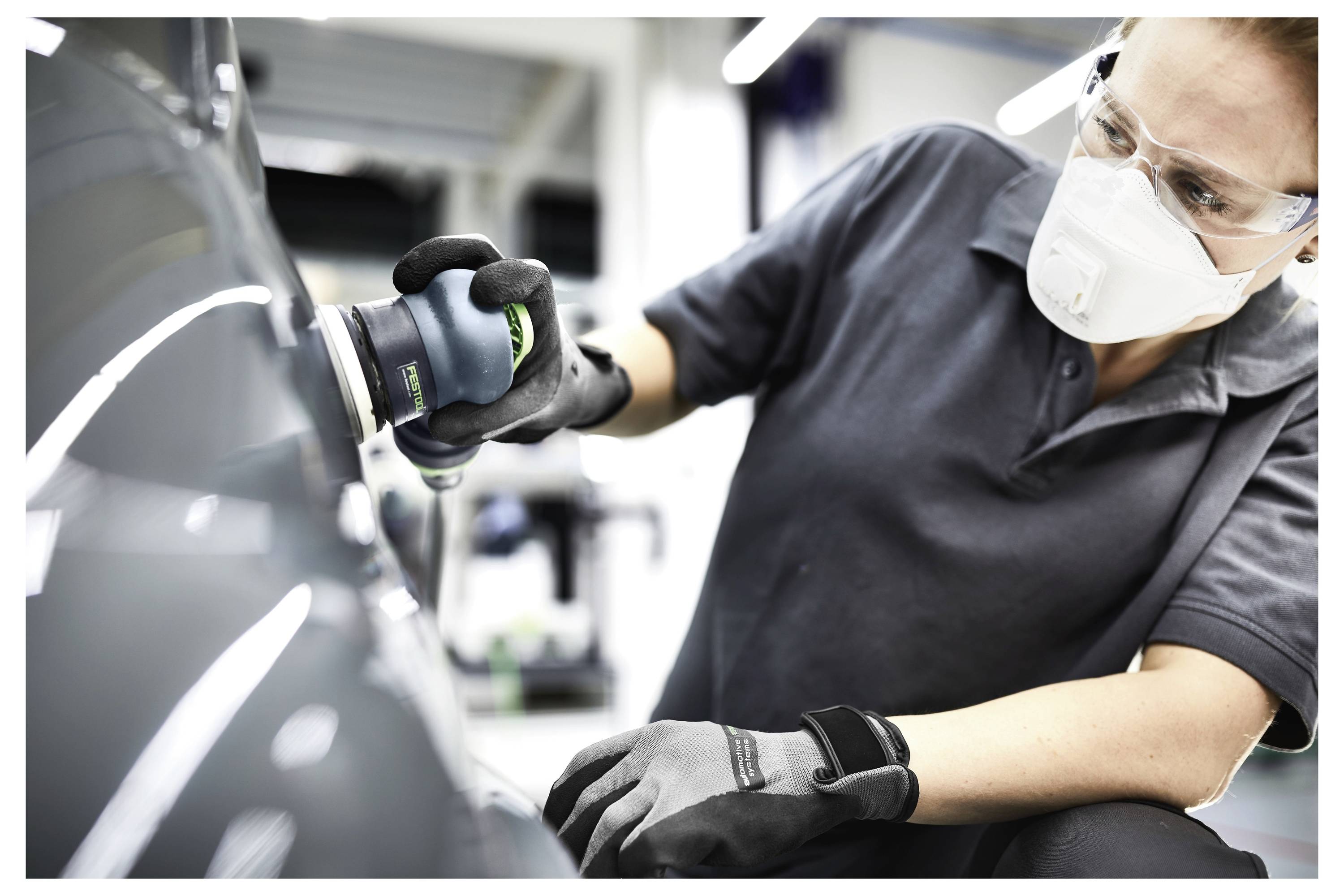A person wearing safety goggles, a mask, and gloves uses a polishing tool on a car's surface in a workshop setting.