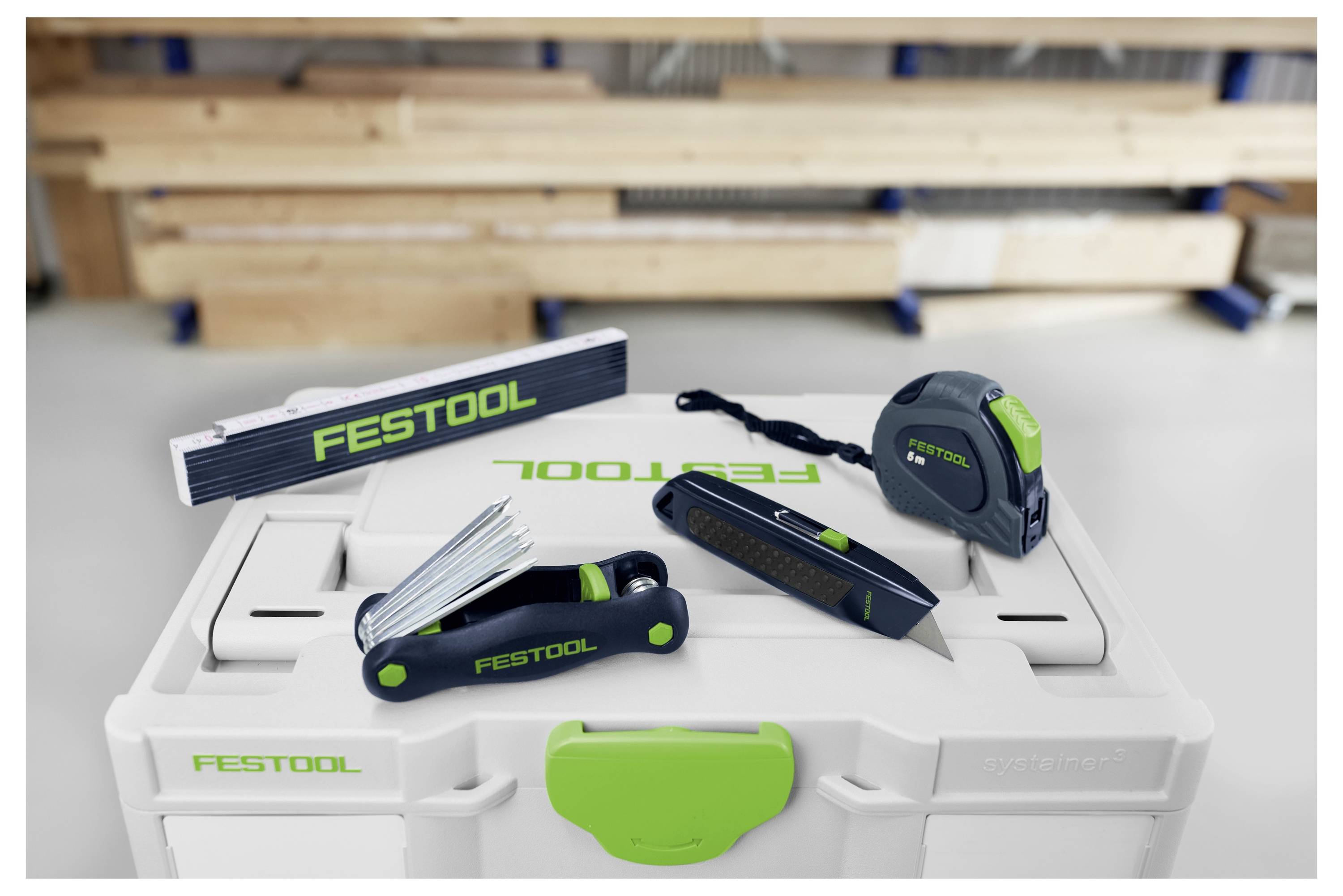 Ein Satz von Festool-Mess- und Schneidwerkzeugen, bestehend aus einem Lineal, einem Stahllineal und einem Universalmesser, auf einem Werkzeugkasten präsentiert.