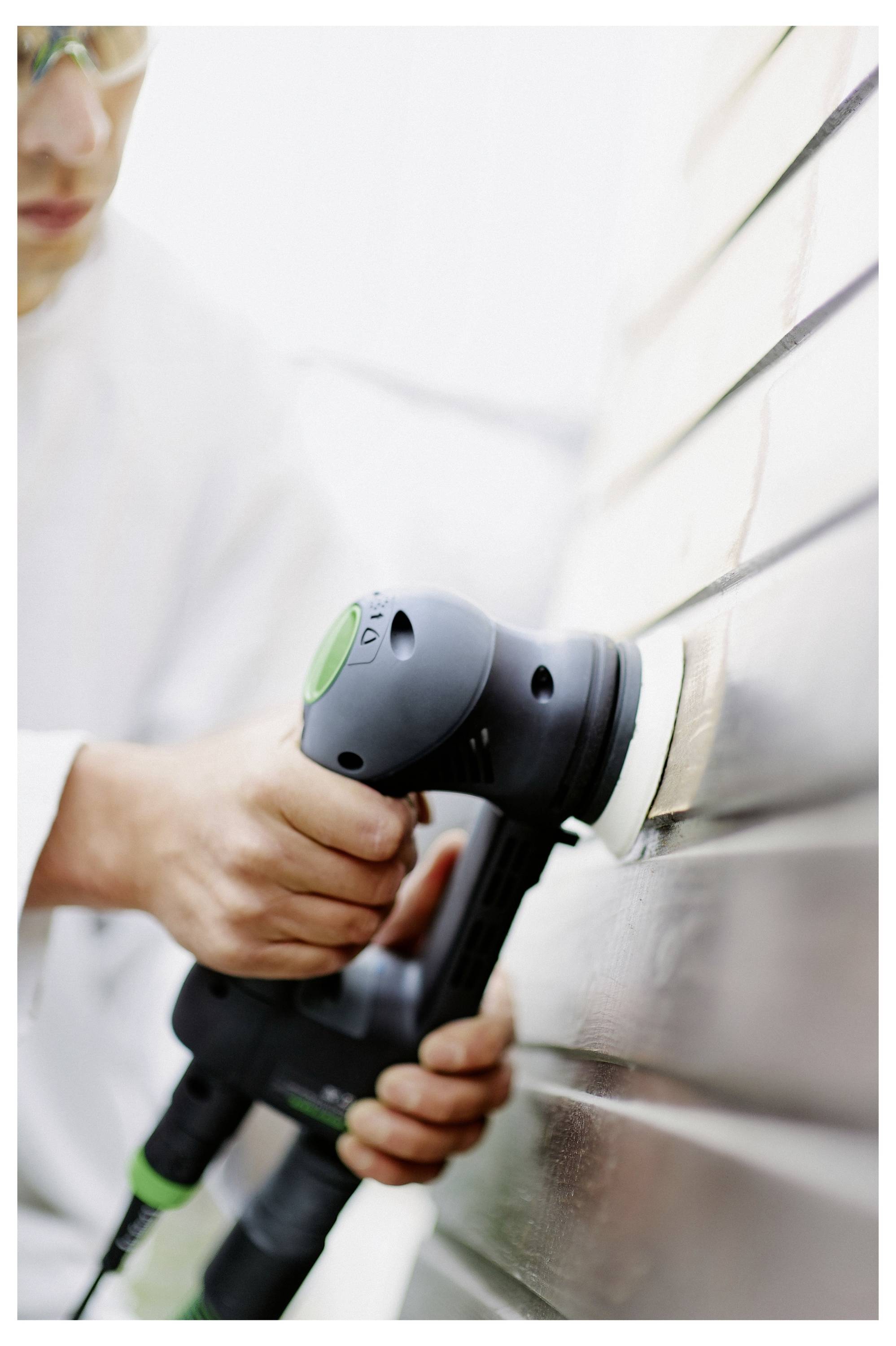 Eine Person, die einen tragbaren elektrischen Schleifers auf einer Holzoberfläche verwendet, um die Bretter zu glätten. Sie trägt eine Schutzbrille und ein weißes Kleidungsstück.