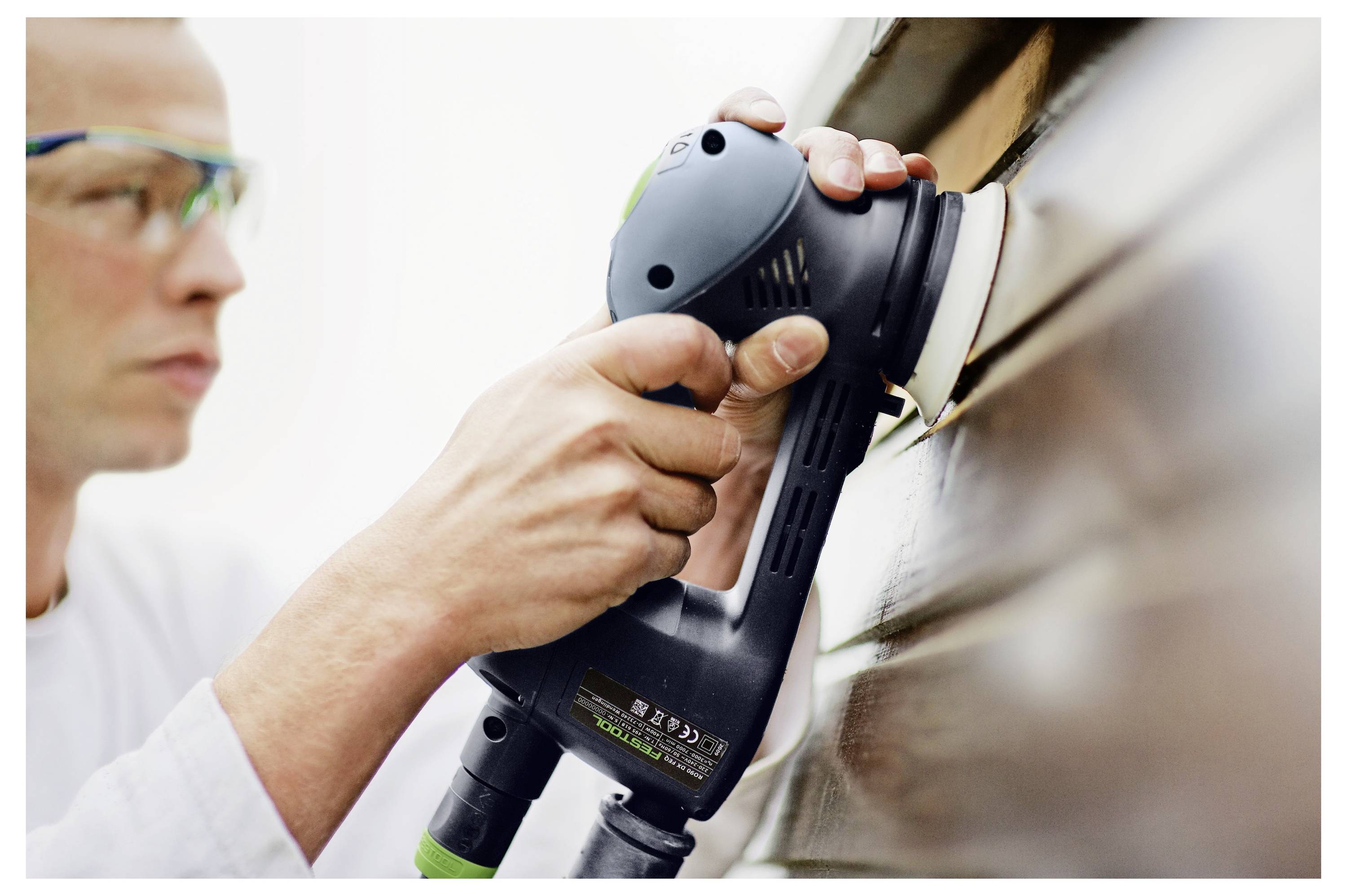 Eine Person, die eine Schutzbrille trägt, verwendet einen elektrischen Schleiffer auf einer Holzverkleidung und konzentriert sich auf das Glätten der Oberfläche.