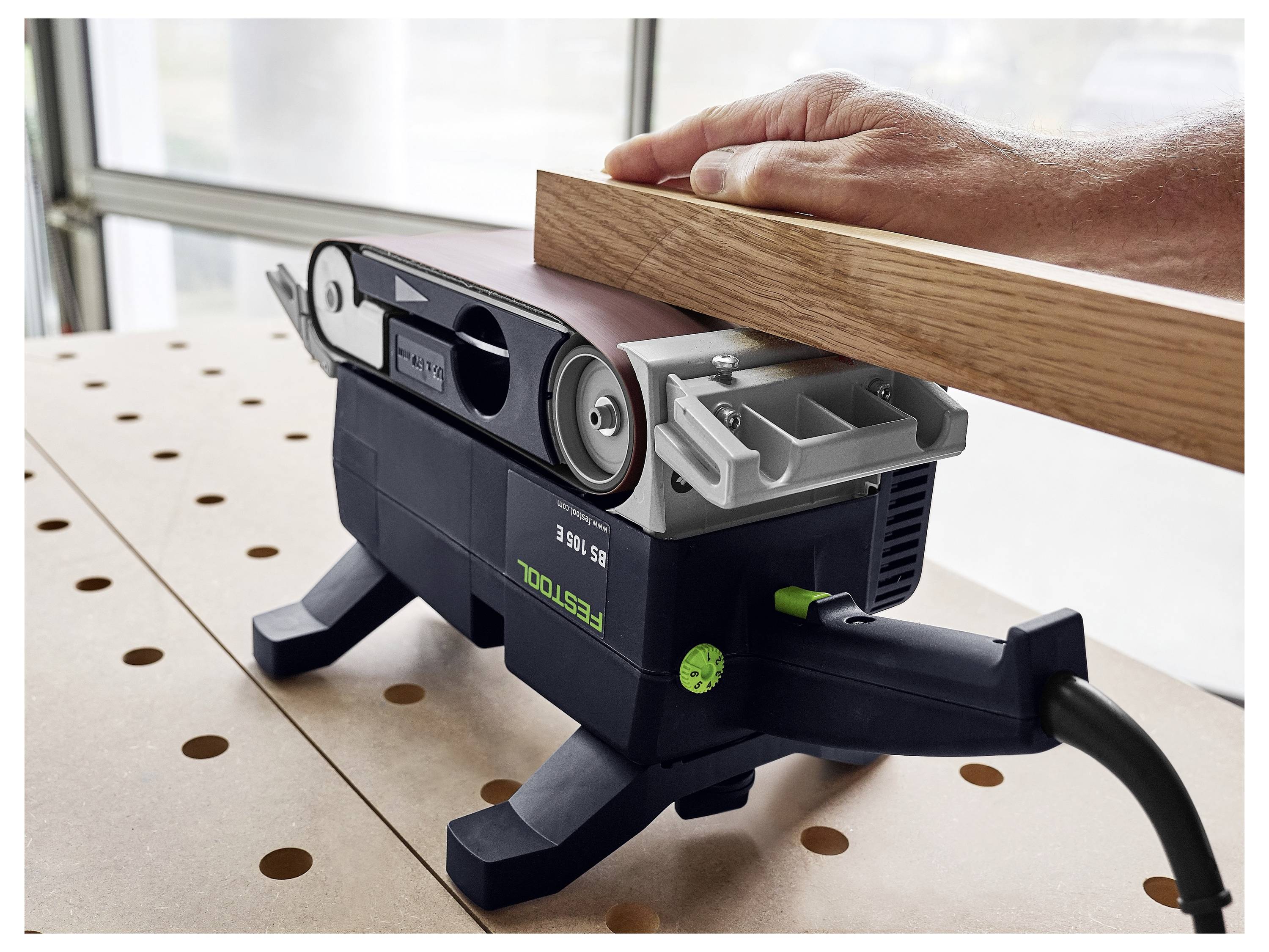 Eine Person schleifen ein Holzbrett mit einem feststehenden Elektroschlifer auf einem Tisch ab. Die Werkstatt ist durch große Fenster gut beleuchtet.