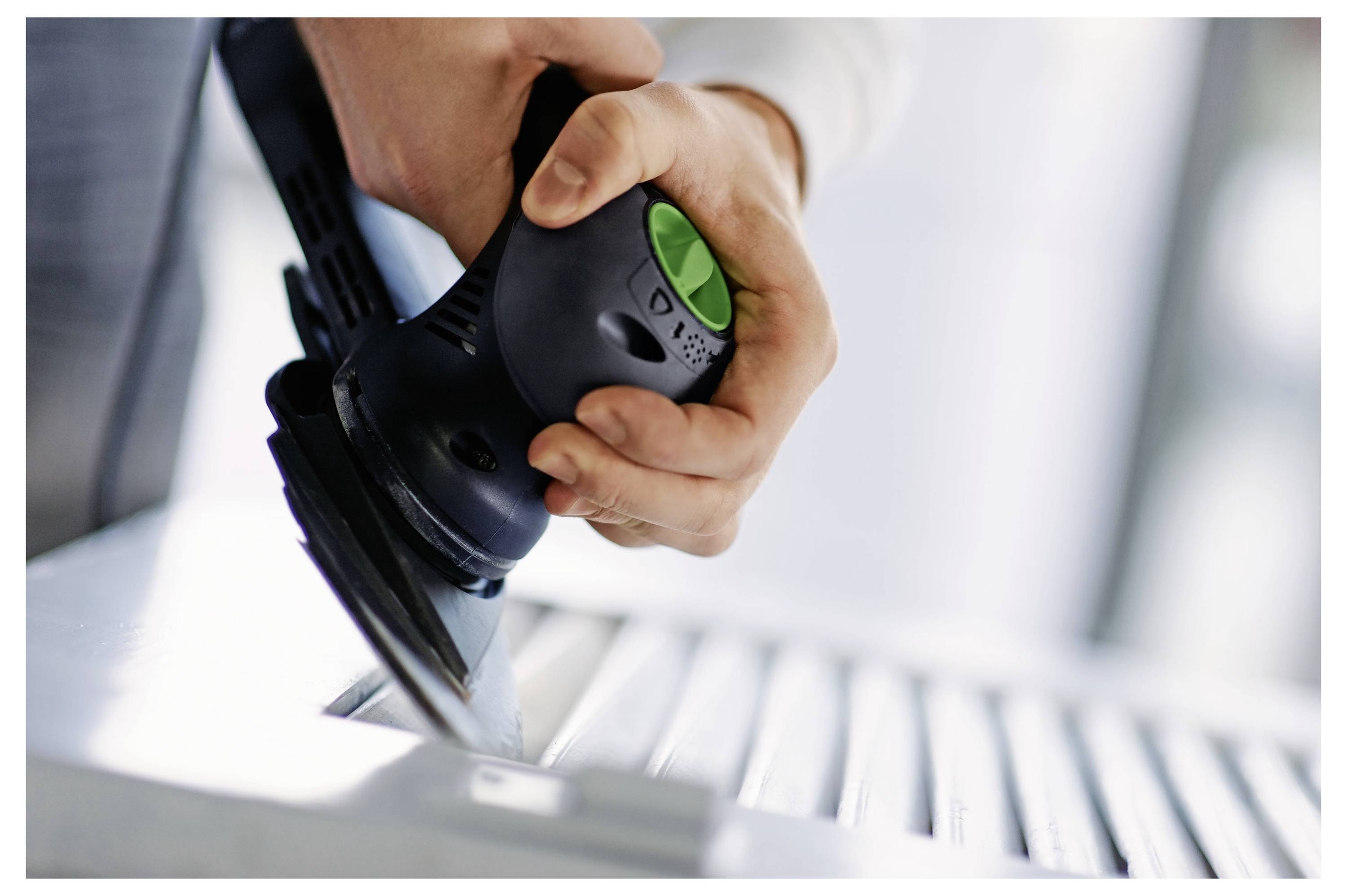 Eine Hand bedient einen kleinen, schwarzgrünen Elektroschleiffer auf einer Holzoberfläche und glättet deren Kanten.