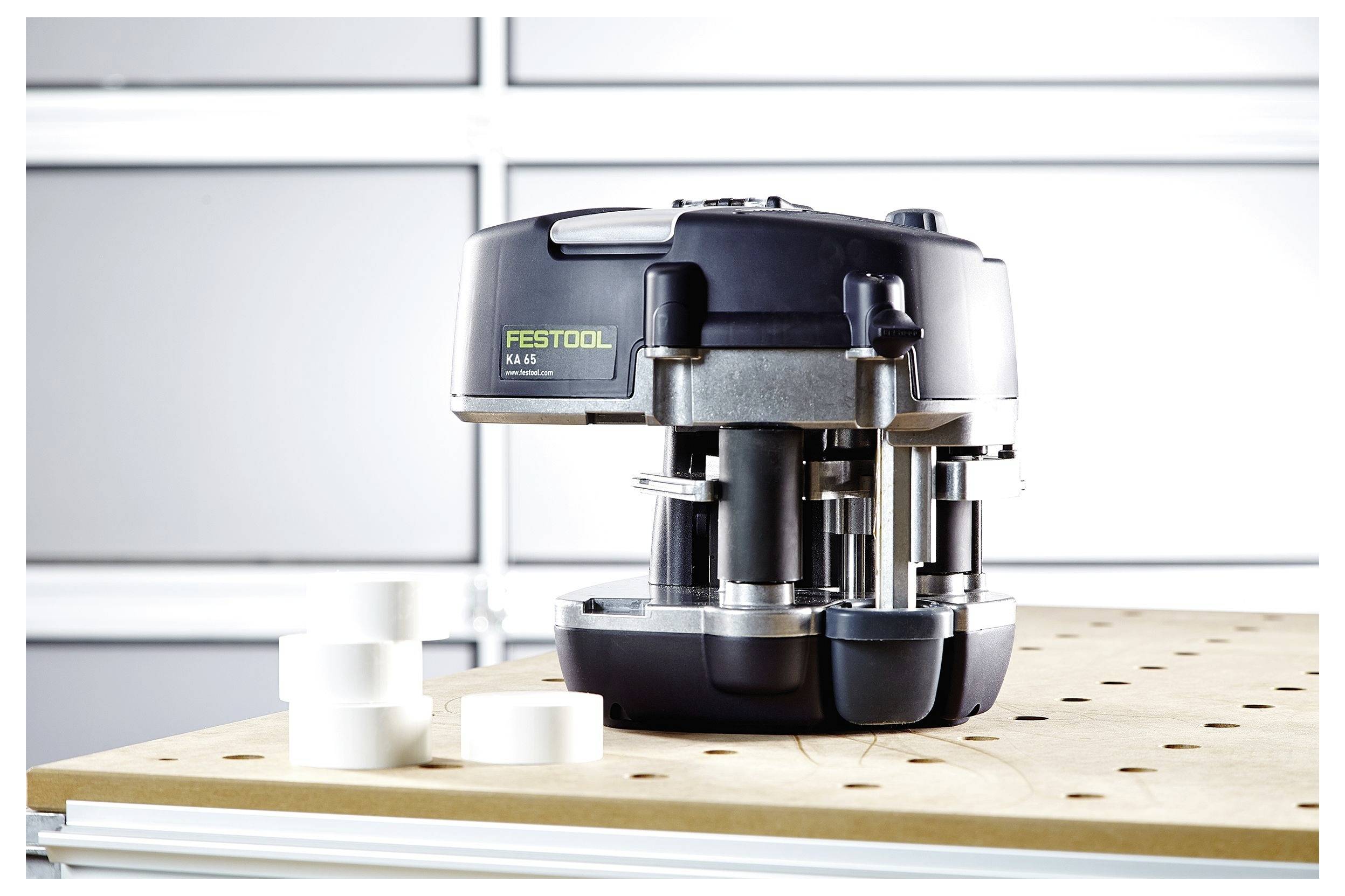 Ein Festool KA 65 Kantenleimgerät steht auf einer perforierten Werkbank, mit mehreren weißen Kantenbandspulen in der Nähe, in einer Werkstattumgebung.
