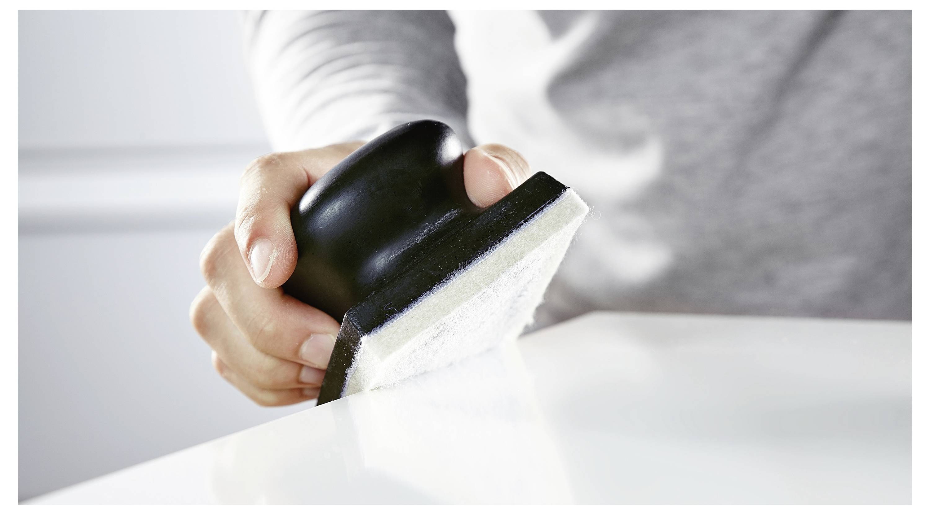Eine Person in einem grauen Hemd benutzt ein Reinigungswerkzeug mit einem schwarzen Griff und einem weißen Tuch, um eine glatte, weiße Oberfläche abzuwischen.