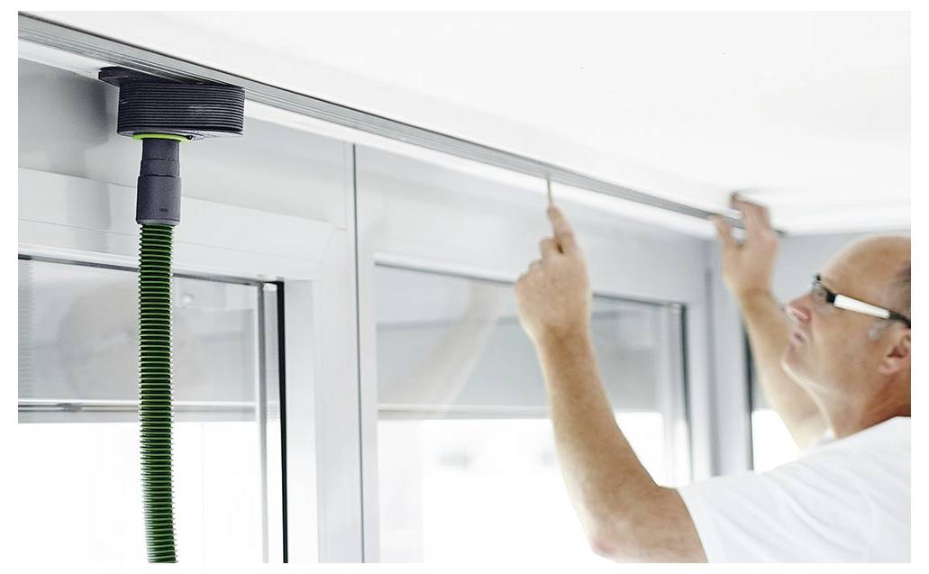 Eine Person misst einen Fensterrahmen mit einem Stahllineal und konzentriert sich auf präzise Anpassung. Ein Werkzeug mit einem grünen Schlauch ist oberhalb des Fensters angebracht.