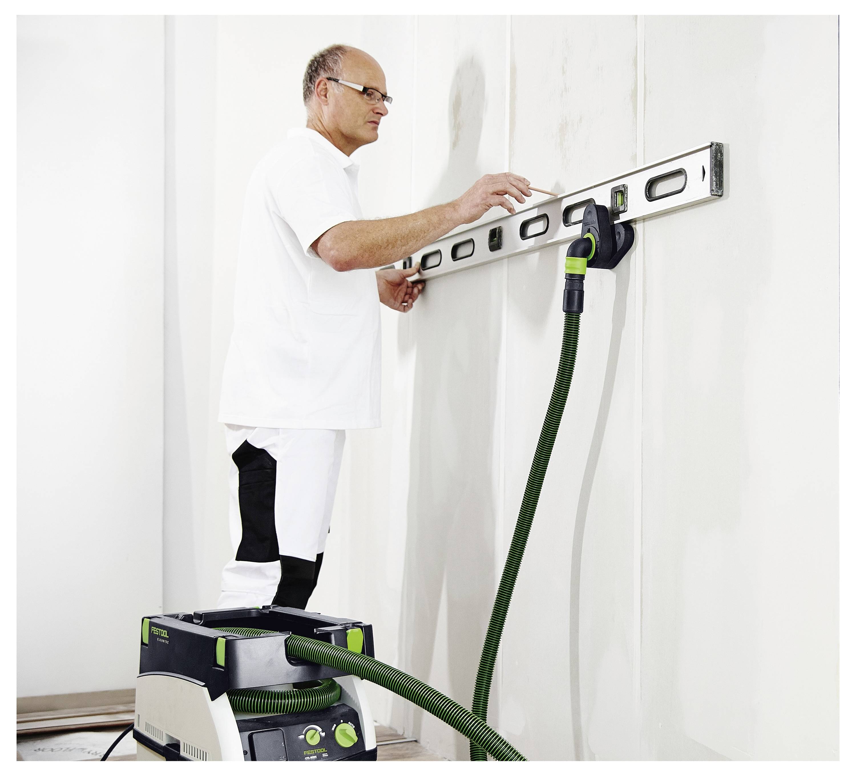 Ein Arbeiter nutzt eine Wasserwaage an einer weißen Wand, während er auf einem Tritthocker steht; ein Staubsauger mit einem grünen Schlauch ist in Benutzung.