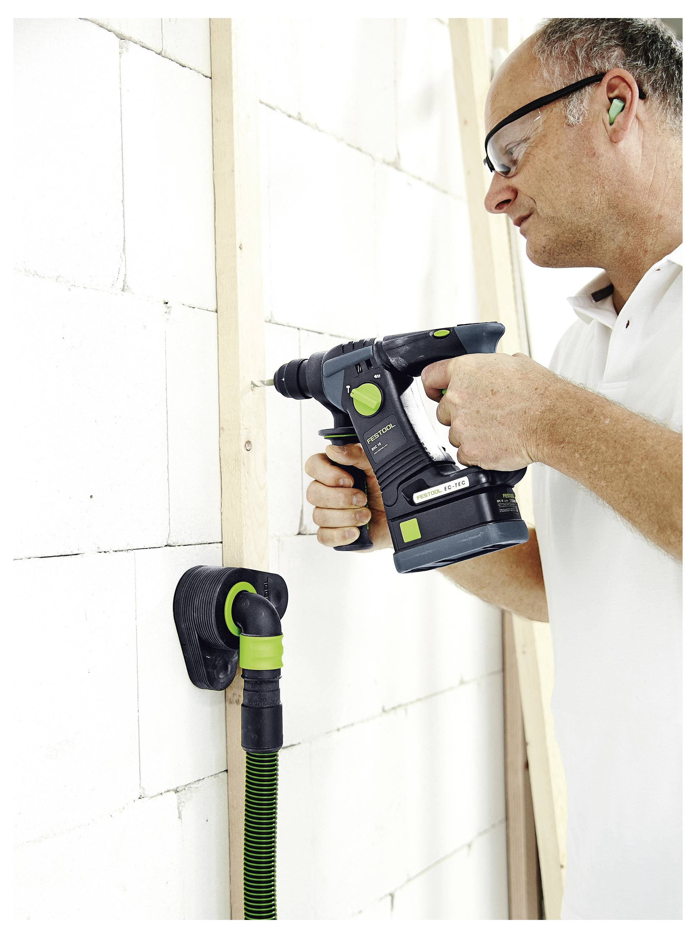 Eine Person, die einen Akku-Bohrer verwendet, um ein Holzbrett an einer weißen Ziegelmauer zu befestigen, während sie eine Schutzbrille und einen Gehörschutz trägt.