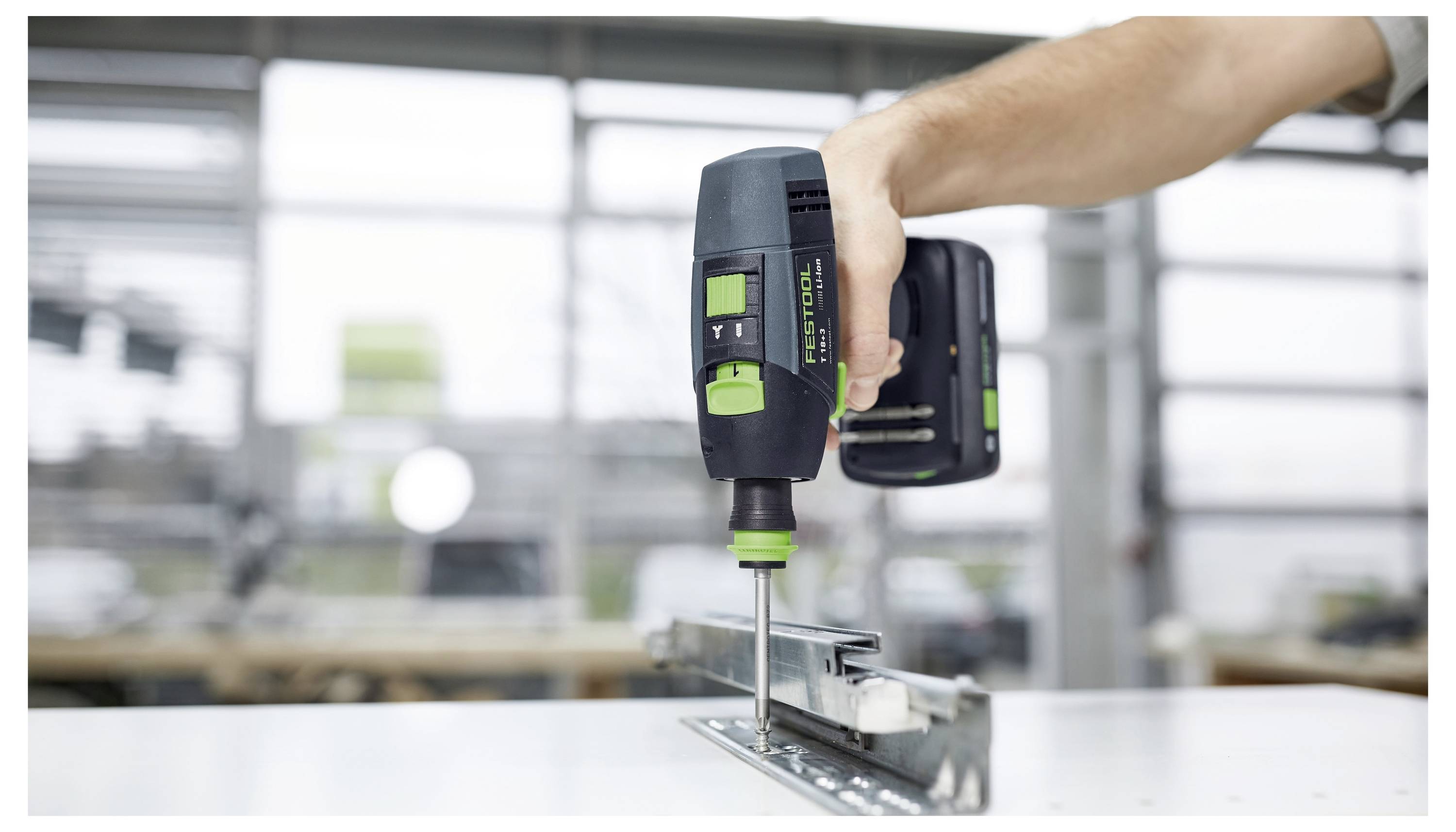 Eine Hand benutzt ein kabelloses Elektrowerkzeug, um in ein Metallscharnier zu bohren, das an einer Oberfläche in einer Werkstattumgebung befestigt ist.