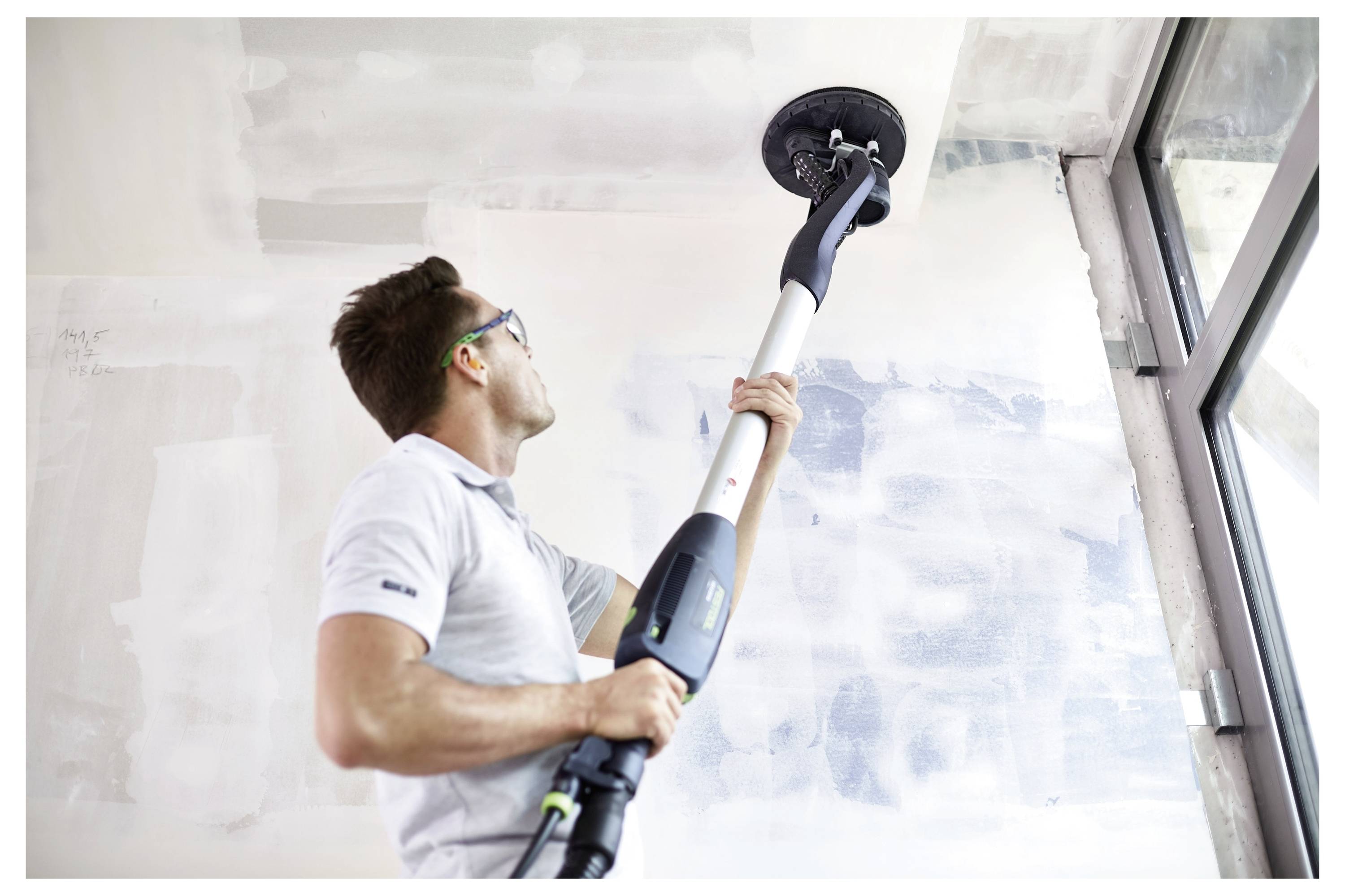 Eine Person mit Schutzbrille benutzt ein Elektrowerkzeug zum Schleifen einer hohen Decke in der Nähe eines großen Fensters und arbeitet in einem renovierungsbedürftigen Raum.
