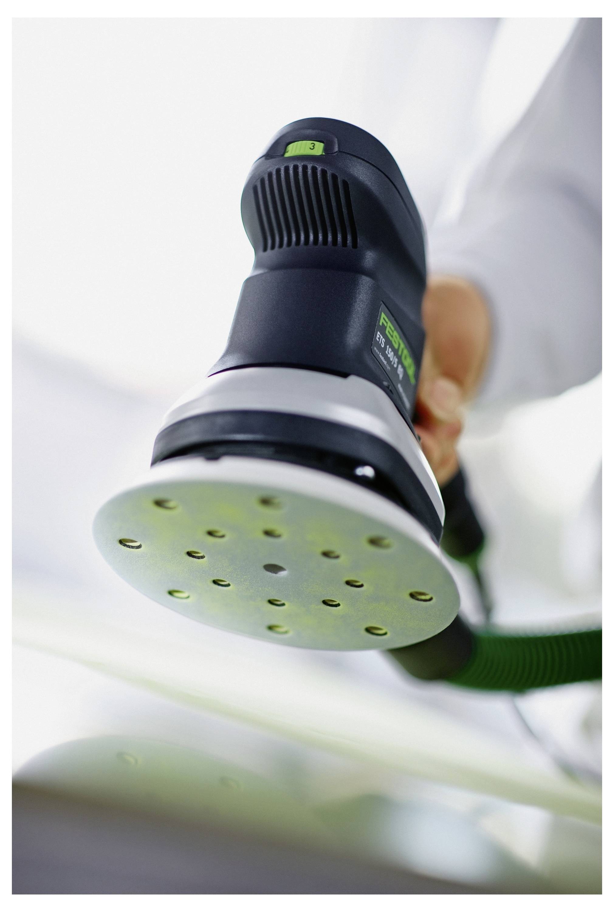 Eine Person in einer Werkstatt bedient eine Elektrosäge und schneidet Holz. Sie trägt eine Schutzbrille und einen Hoodie. Im Hintergrund sind Holzstapel sichtbar.