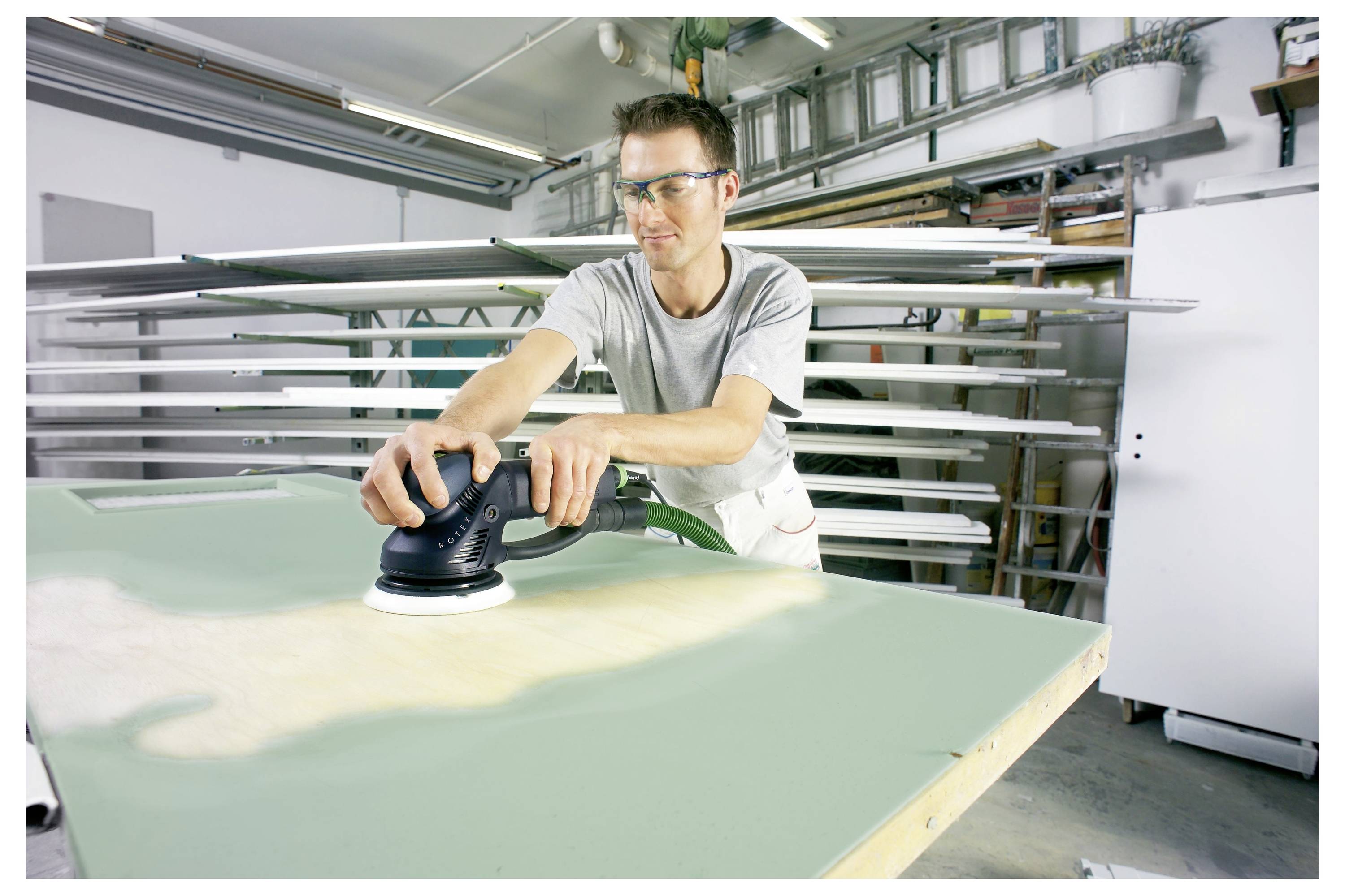 Eine Person, die eine Schutzbrille trägt, verwendet einen elektrischen Schleiffer auf einem grünen Brett in einer Werkstatt, umgeben von Metallstangen und Regalen.