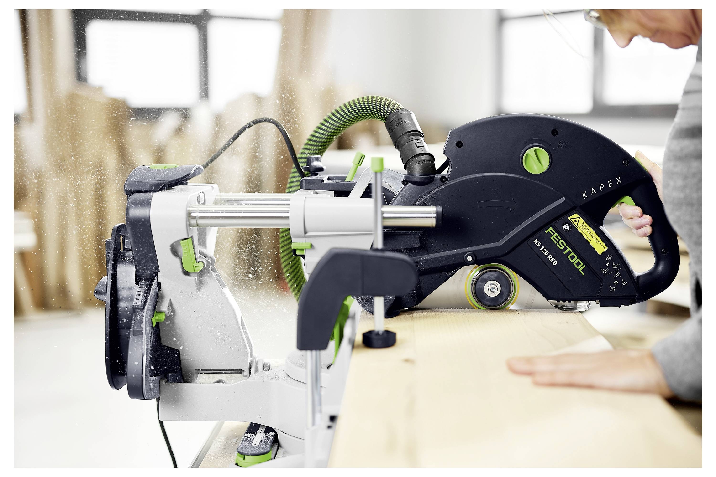 Eine Person benutzt eine Festool Kapex Säge, um ein Holzbrett zu schneiden, mit Sägespänen in der Luft, in einer Werkstattsumgebung.