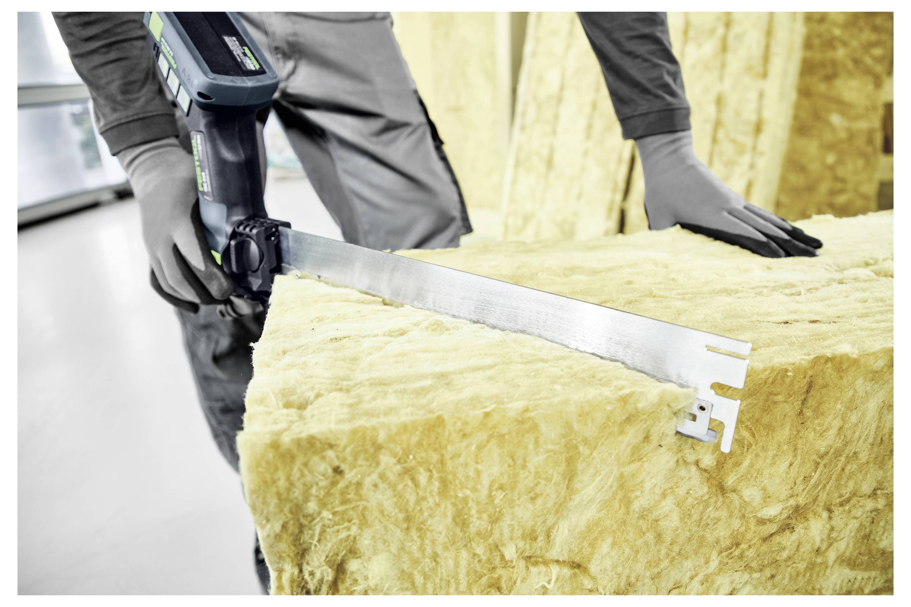 Eine Person, die Handschuhe trägt, verwendet eine elektrische Säge, um einen großen Block aus Isoliermaterial, wahrscheinlich Glasfaser oder Mineralwolle, in einer Werkstatt zu schneiden.