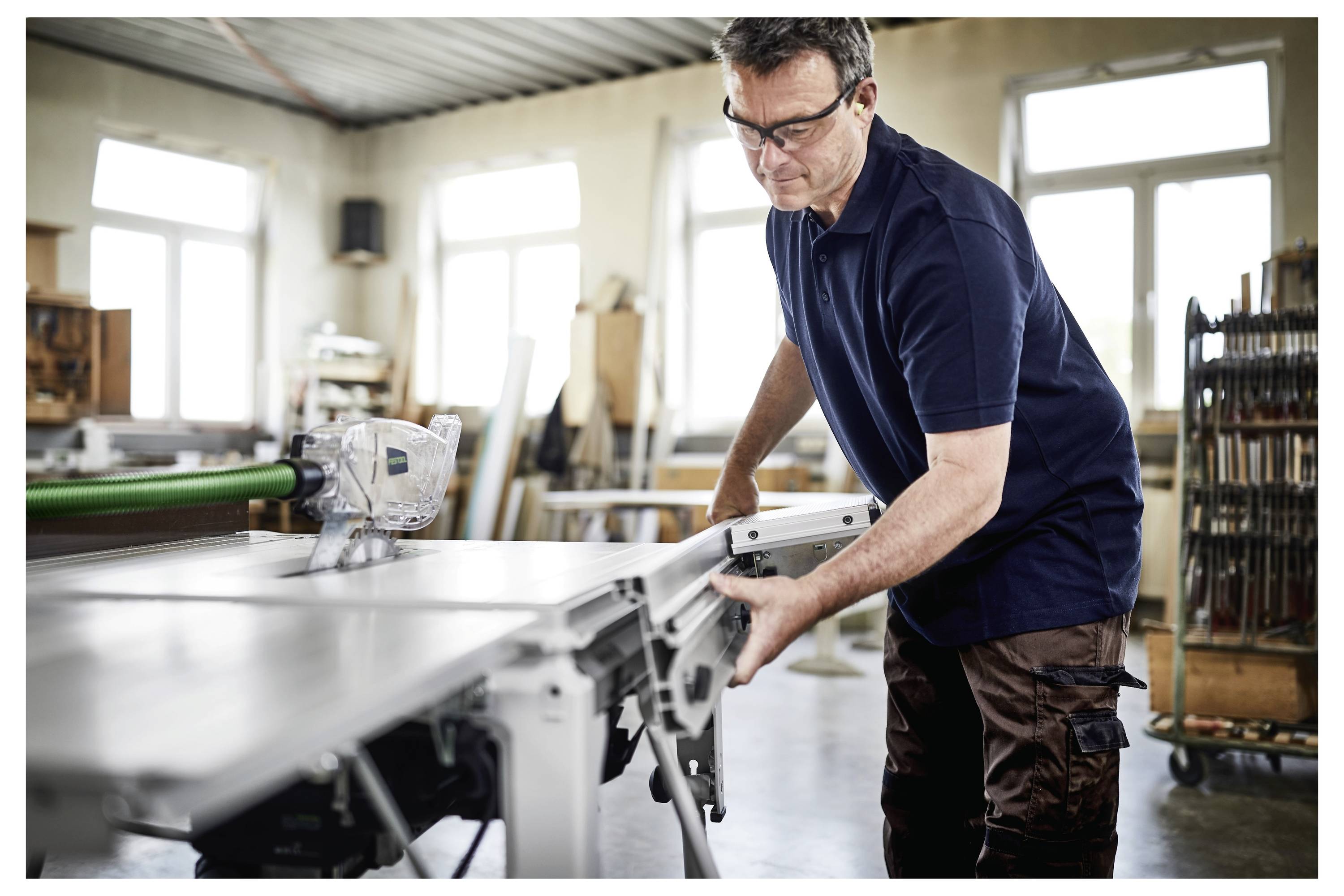 Ein Mann mit Schutzbrille bedient eine Tischkreissäge in einer Werkstatt und konzentriert sich auf seine Arbeit, während verschiedene Werkzeuge und Materialien um ihn herum sichtbar sind.