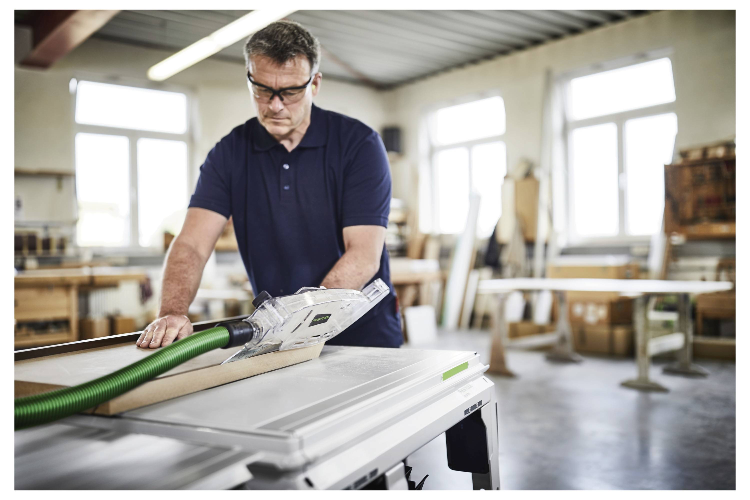 Eine Person mit Schutzbrille bedient eine Tischkreissäge in einer gut beleuchteten Werkstatt und glättet ein Stück Holz.