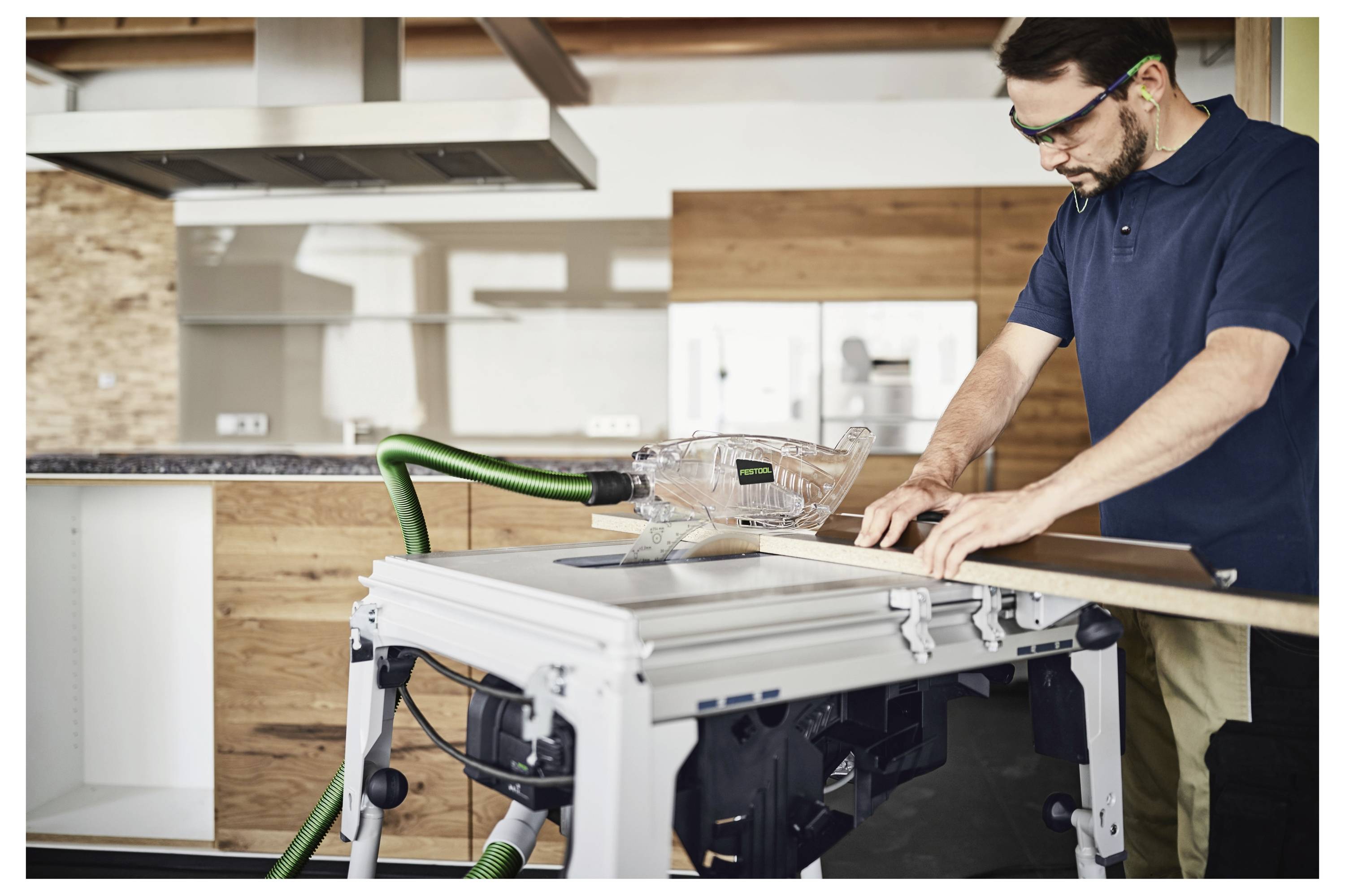 Eine Person, die eine Schutzbrille und einen Gehörschutz trägt, bedient eine Tischkreissäge auf einer Holzplatte in einer modernen Werkstattumgebung.