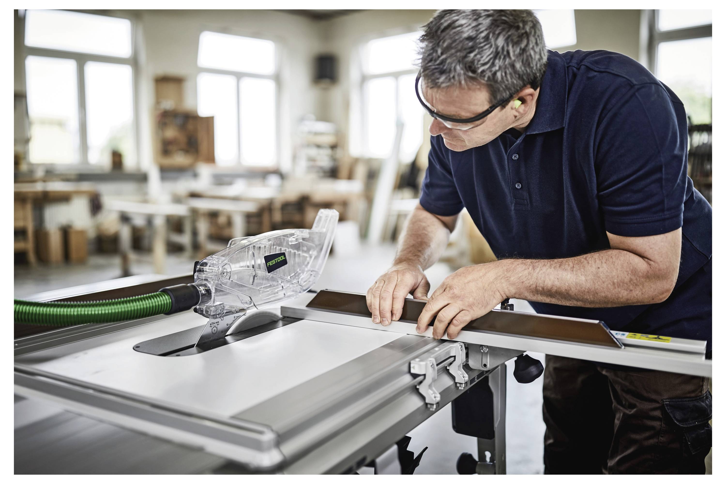 Ein Mann mit Schutzausrüstung bedient eine Tischkreissäge und schneidet ein Stück Holz. Im Hintergrund sind eine Werkstatt mit Ausrüstung und Schränken zu sehen.