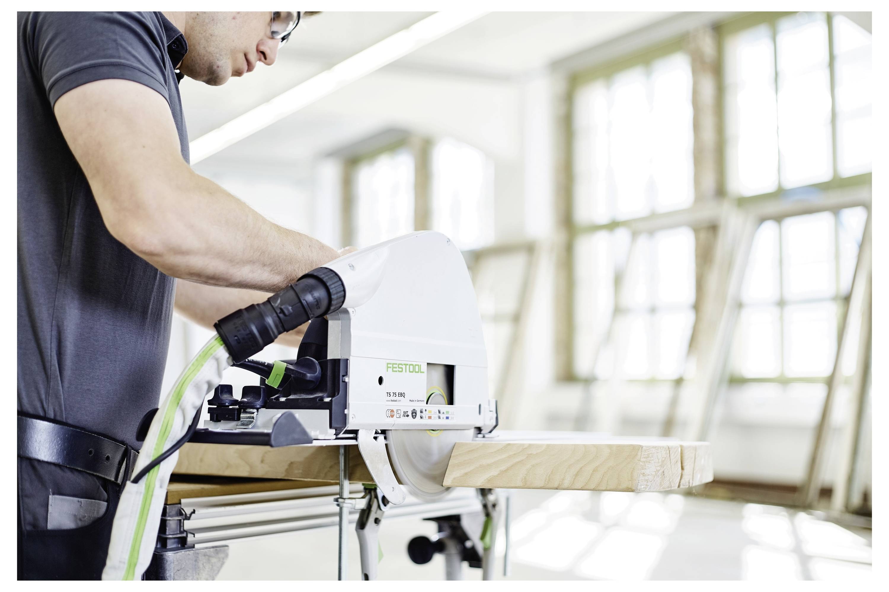 Eine Person bedient eine Kreissäge auf einem Holzbrett in einer hellen, modernen Werkstatt mit großen Fenstern.