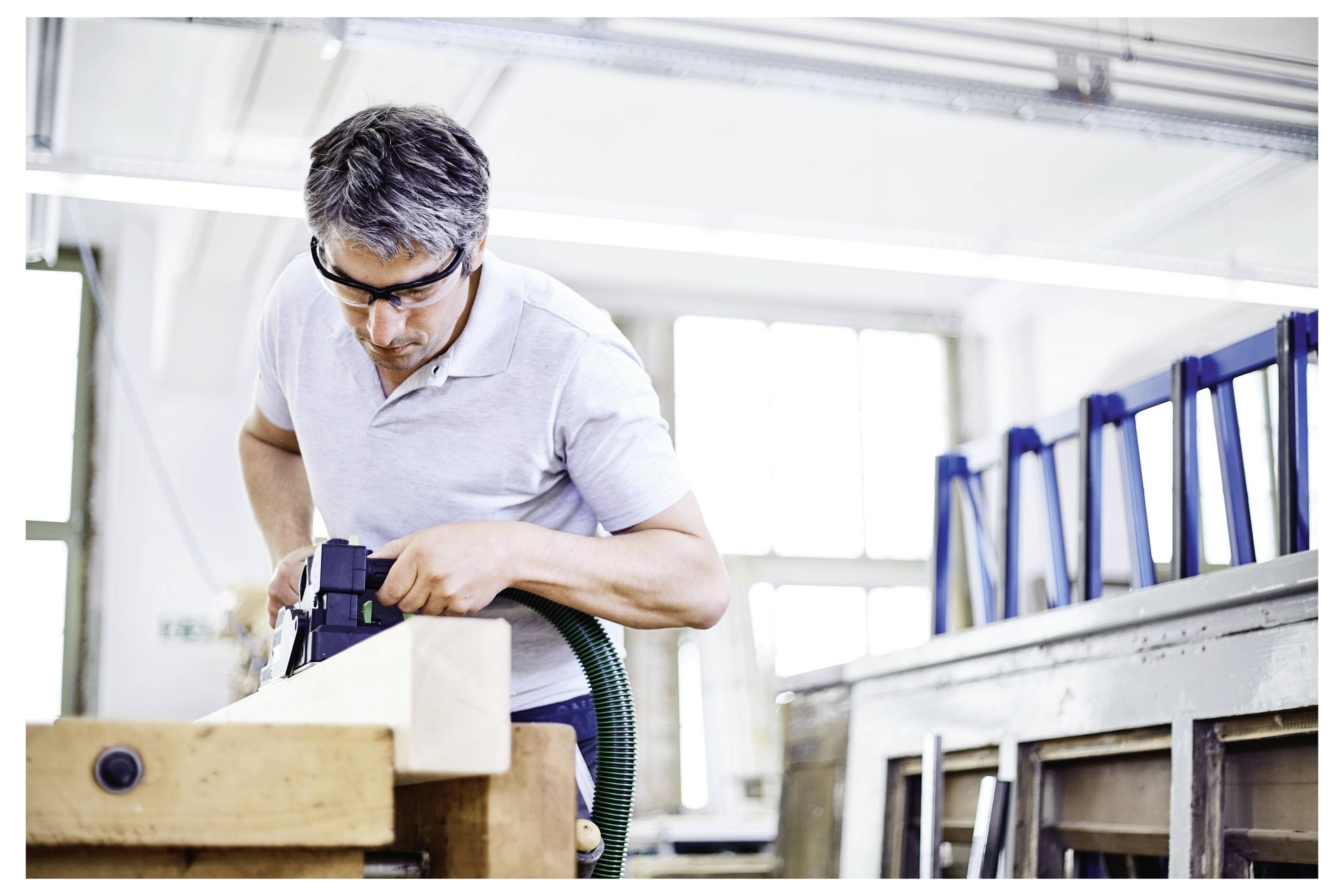 Eine Person in Schutzbrille und grauem Hemd verwendet ein Elektrowerkzeug an einem Holzstück in einer gut beleuchteten Werkstatt.