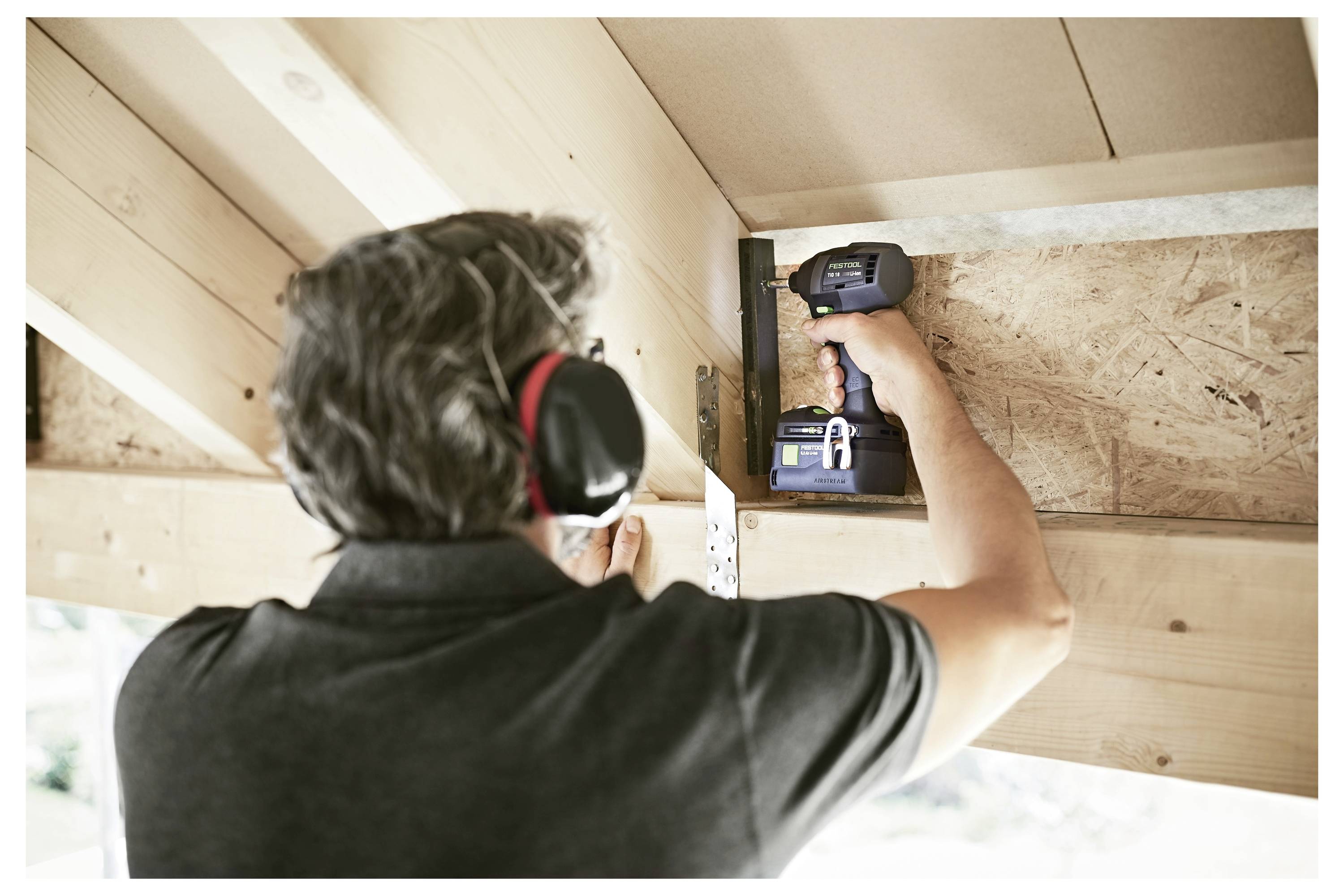 Eine Person mit Gehörschutz benutzt einen Elektroschrauber, um einen Holzbalken unter einer Decke zu befestigen, was auf Bau- oder Renovierungsarbeiten hindeutet.