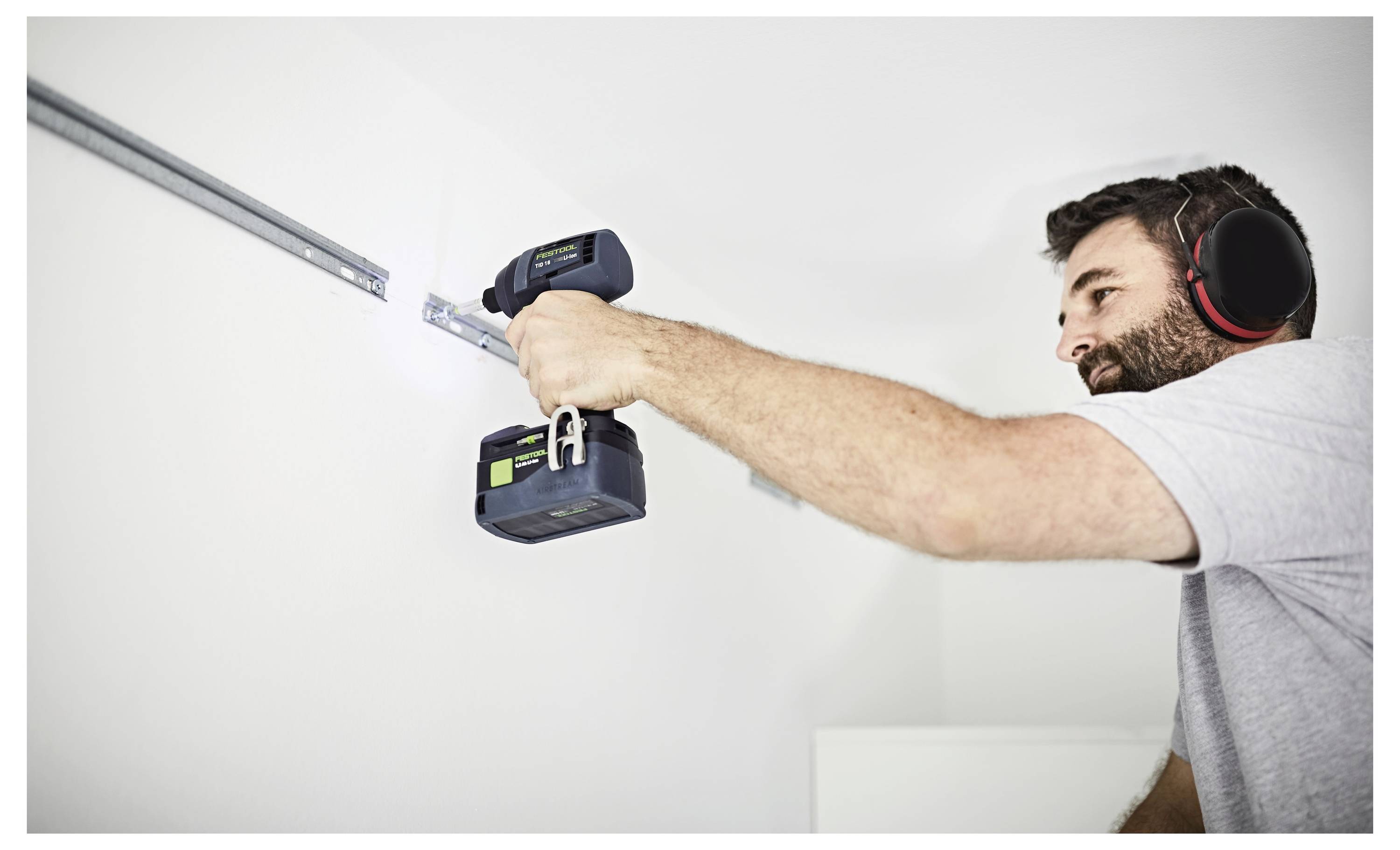 Eine Person in einem grauen Hemd benutzt eine Elektrobohrmaschine, um eine Metallschiene an einer weißen Wand zu montieren. Sie trägt zum Schutz einen Gehörschutz.