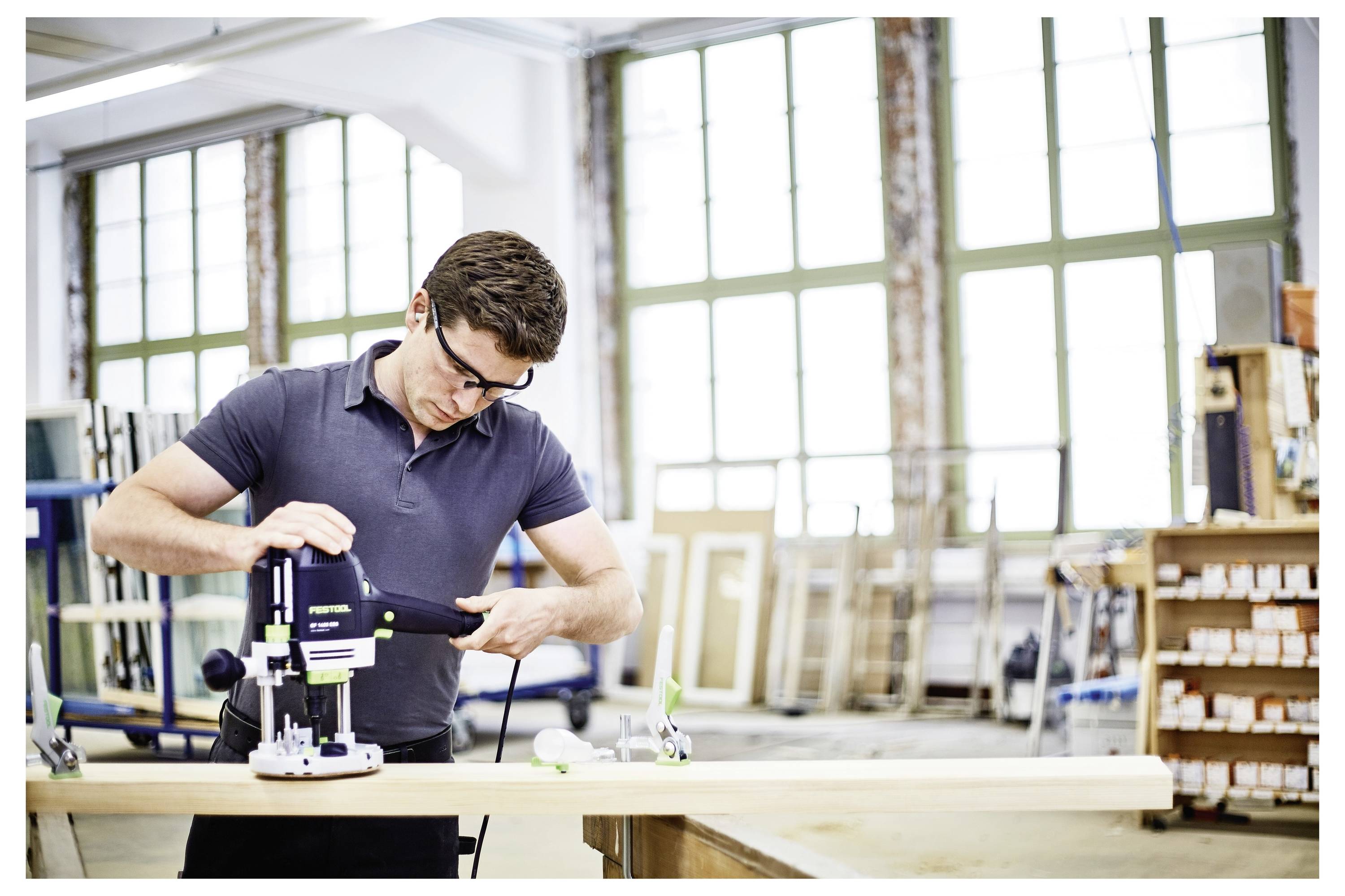 Eine Person in einer Schutzbrille bedient ein Elektrowerkzeug auf einer Holzplatte in einer hellen Werkstatt, die von großen Fenstern umgeben ist und verschiedene Werkzeuge enthält.