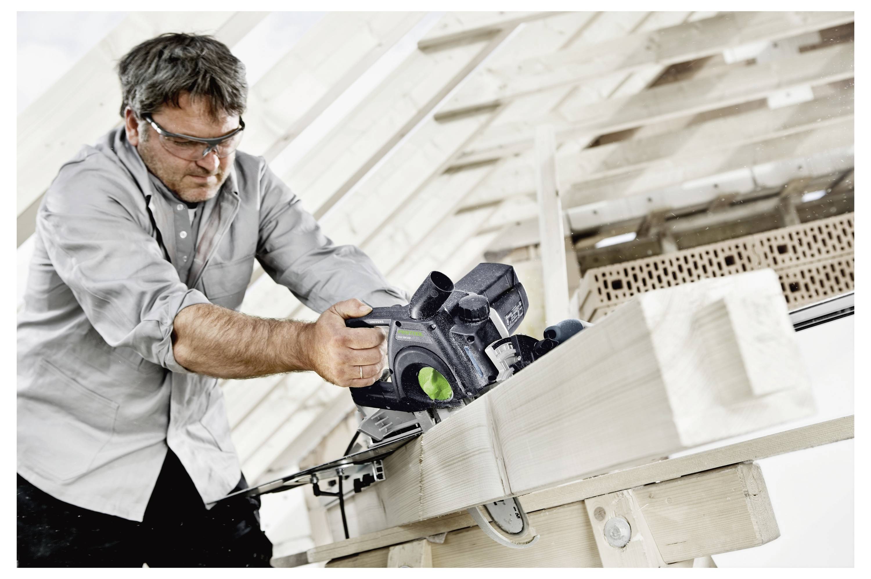 Ein Mann mit einer Schutzbrille benutzt eine Kreissäge, um einen Holzbalken in einem teilweise konstruierten Gebäude zu schneiden.
