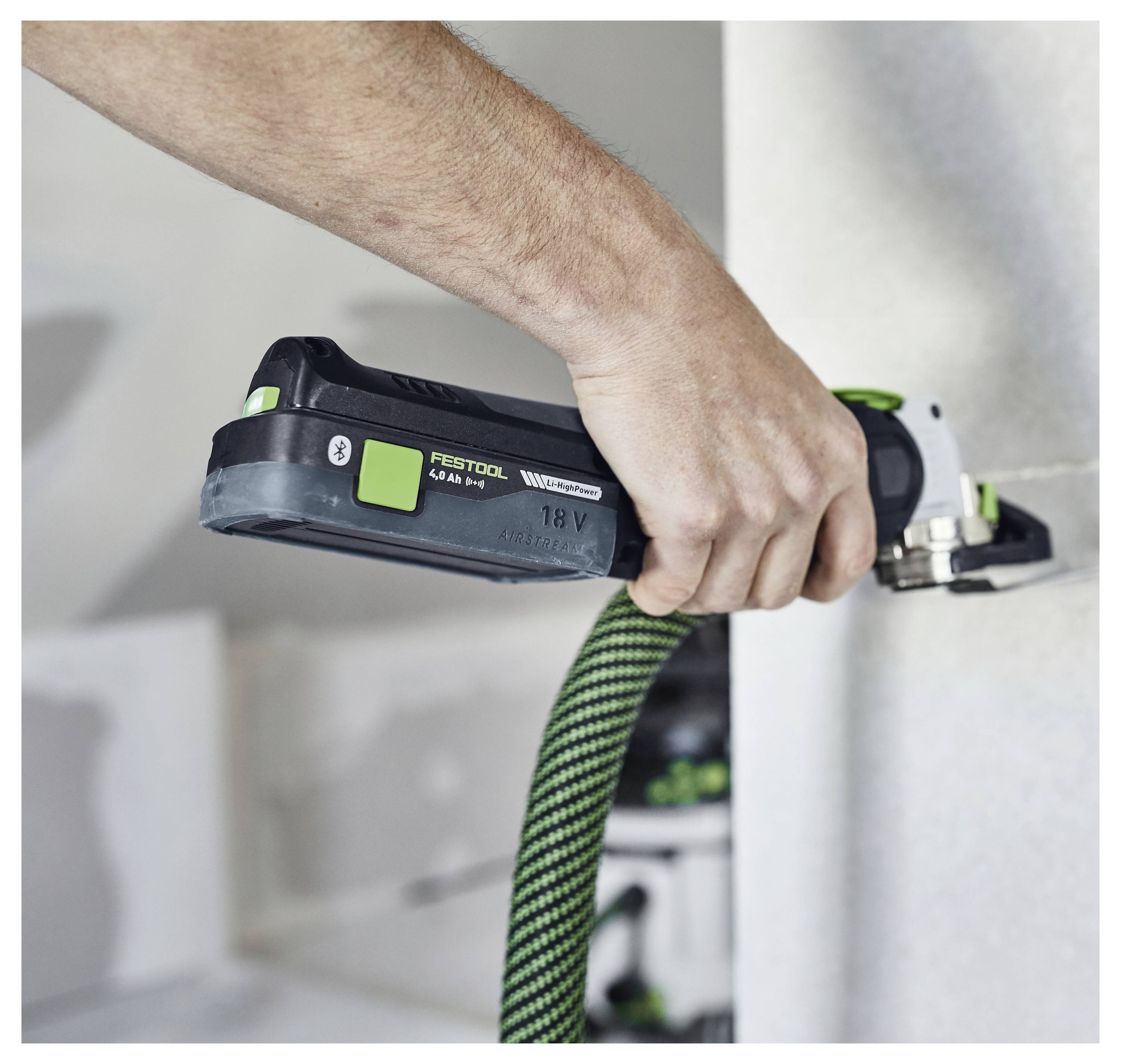 Eine Person benutzt ein elektrisches Elektrowerkzeug mit einem grün-schwarzen Griff, um eine Trockenbauwand zu glätten. Das Werkzeug ist mit einem Schlauch verbunden.