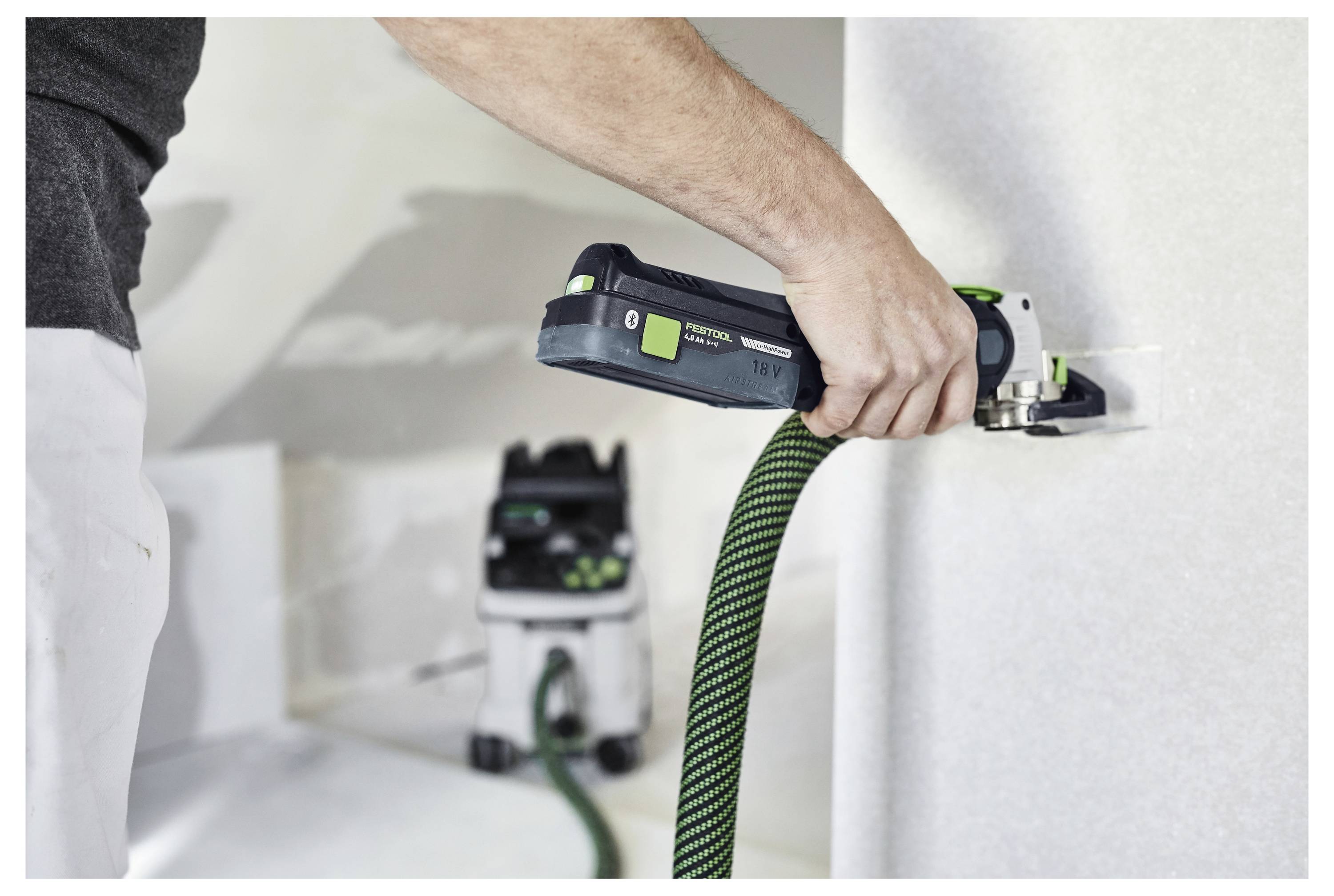 Eine Person benutzt ein Elektrowerkzeug, das mit einem Staubsaugsystem verbunden ist, um Staub zu absaugen, während sie an einer Wand arbeitet.