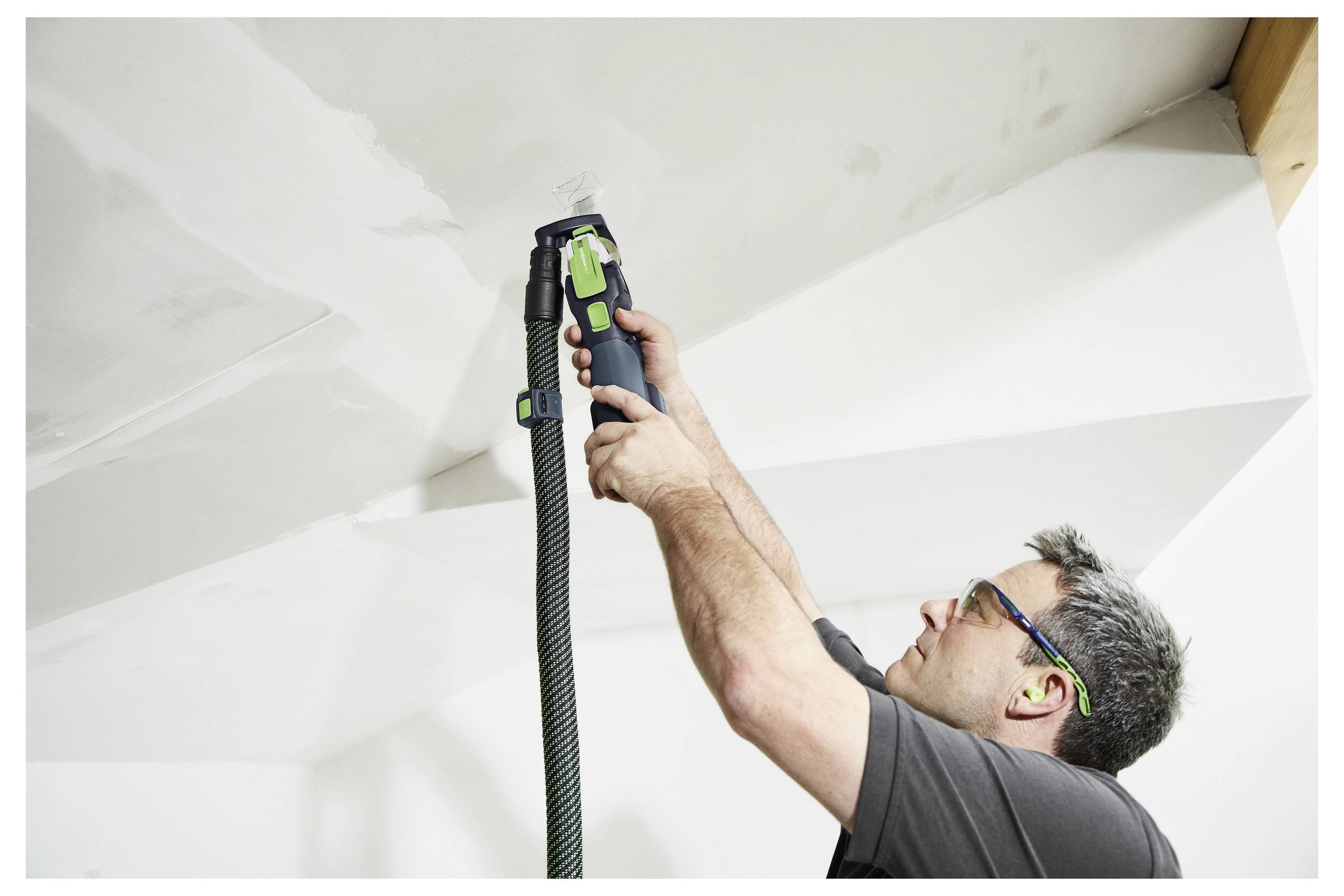 Eine Person in Schutzbrille und Gehörschutz schleifen eine weiße Decke mit einem Elektrowerkzeug mit angebautem Staubsammler.