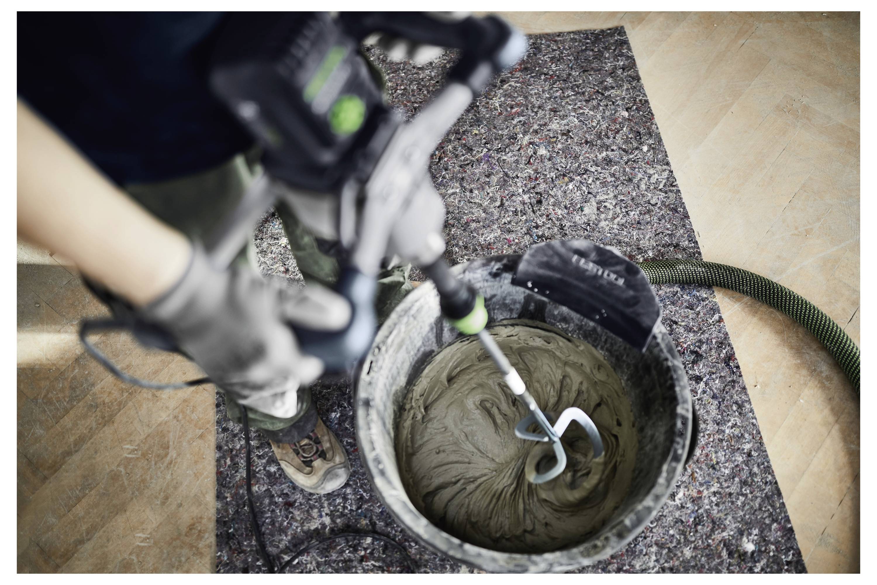 Eine Person benutzt einen Elektroschrauber mit einem Rühraufsatz, um Material in einem Eimer zu rühren, wahrscheinlich zur Vorbereitung von Baumaterial für eine Bauaufgabe.