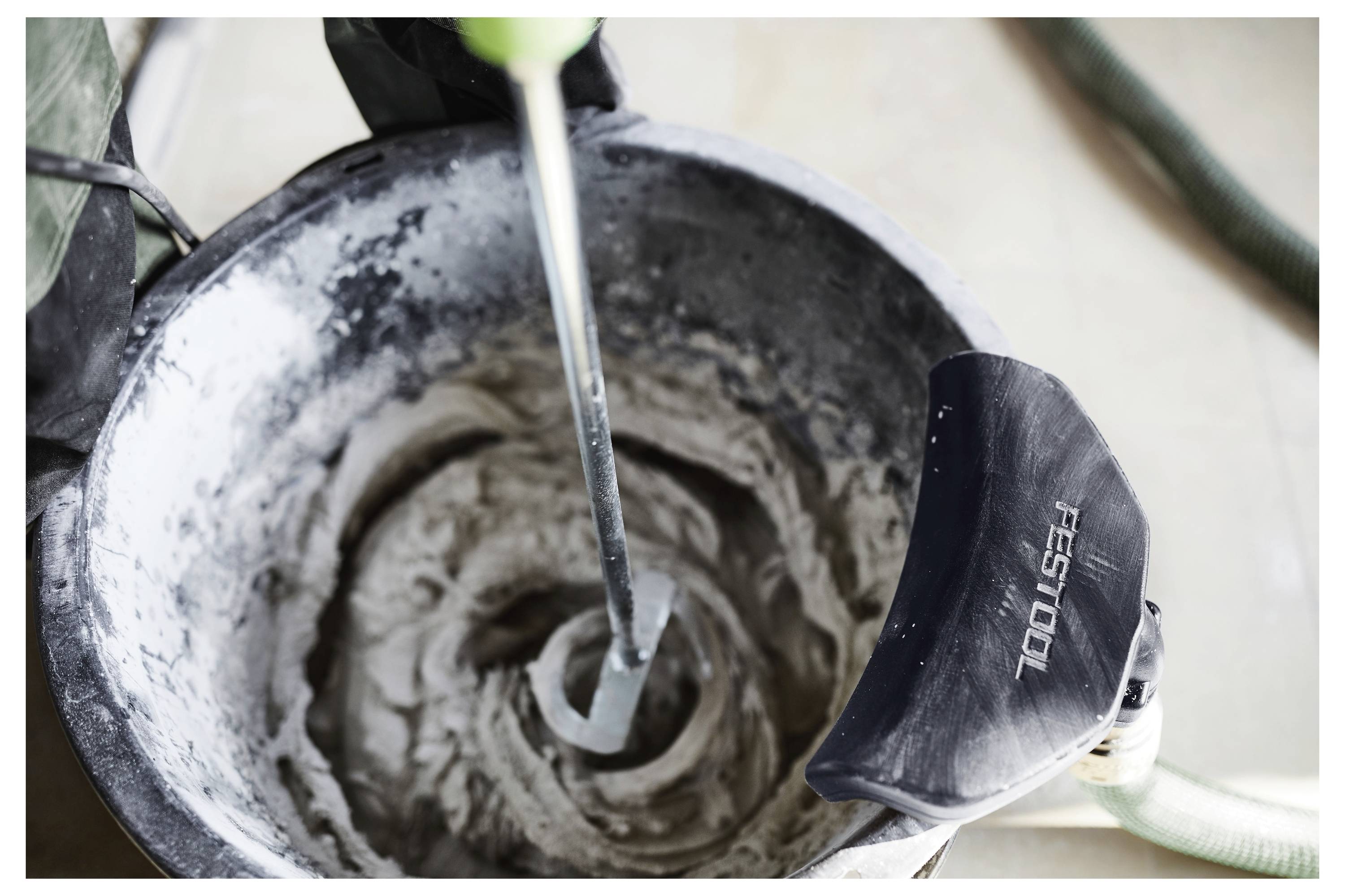 Ein Werkzeug mischt grauen Zement in einem Eimer und zeigt eine Draufsicht des wirbelnden Gemisches mit einem schwarzen Festool-Staubsaugeransatz an der Seite.