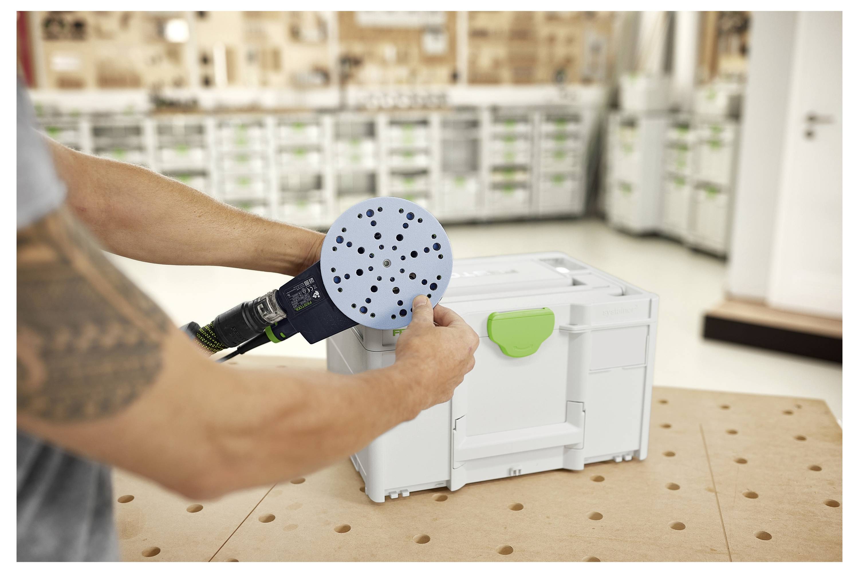 Eine Person stellt eine runde Schleifscheibe auf einem Elektroschleiffer über einer Holzwerkbank ein. Eine große Lagerkiste befindet sich im Hintergrund.