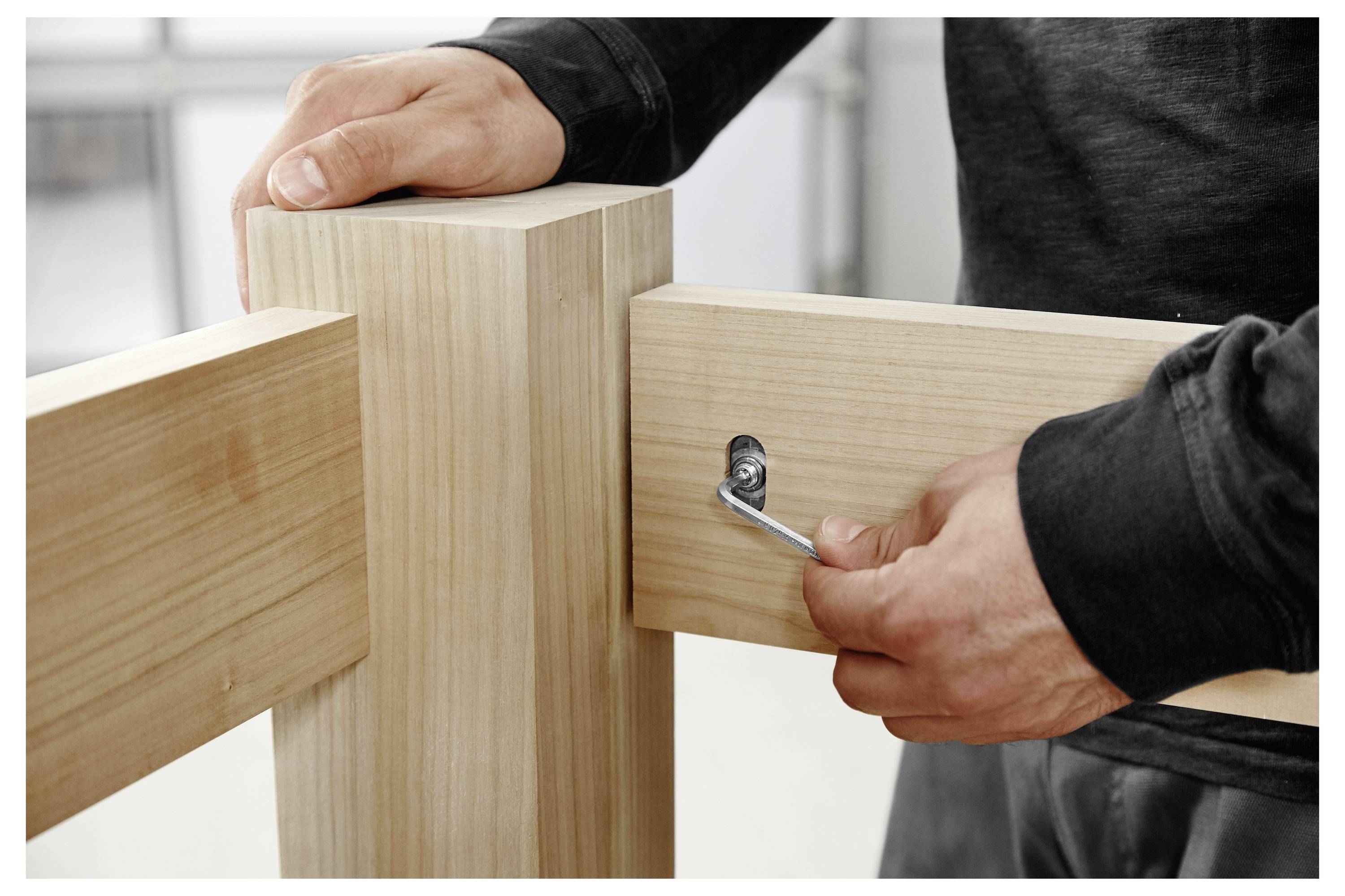 Eine Person zieht eine Schraube mit einem Innensechskantschlüssel an und verbindet dabei zwei Holzbalken im rechten Winkel.