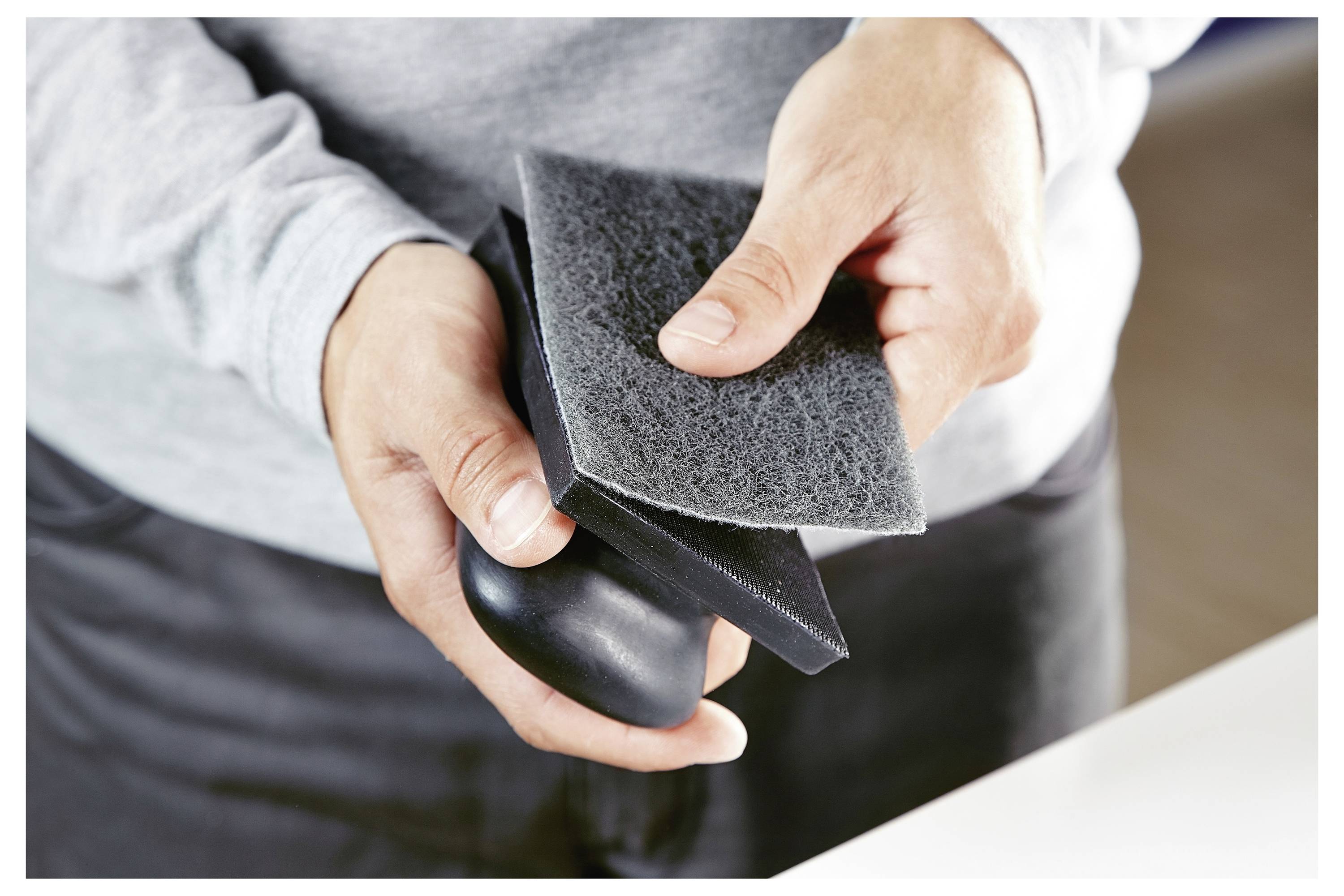 Hände halten ein graues Scheuertuch und einen schwarzen Gegenstand und scheinen eine Reinigungs- oder Polierbewegung zu demonstrieren.