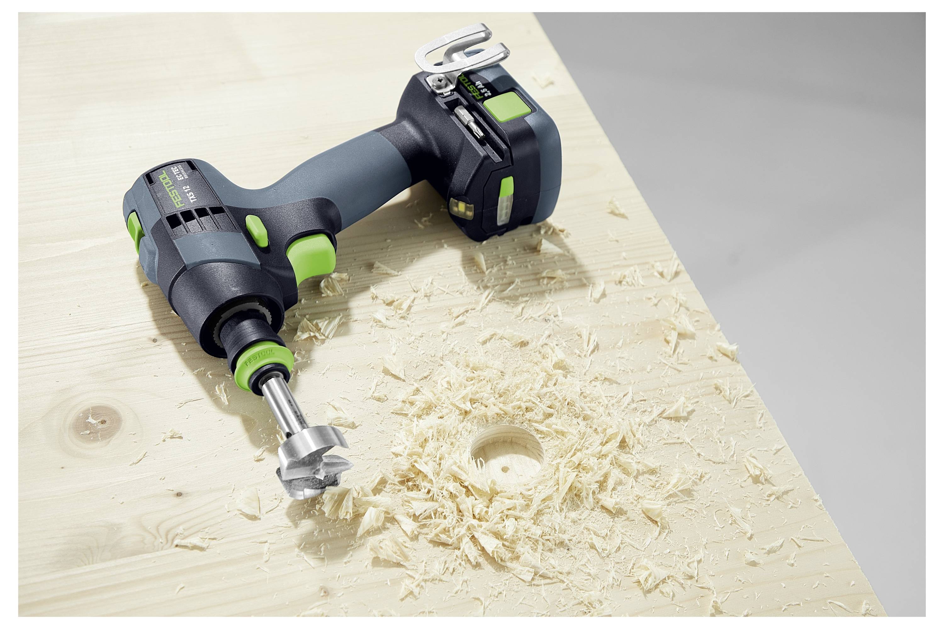 Ein Elektrobohrer mit angebrachtem Bohrer liegt auf einer hellen Holzoberfläche mit Holzspänen rund um ein gebohrtes Loch.