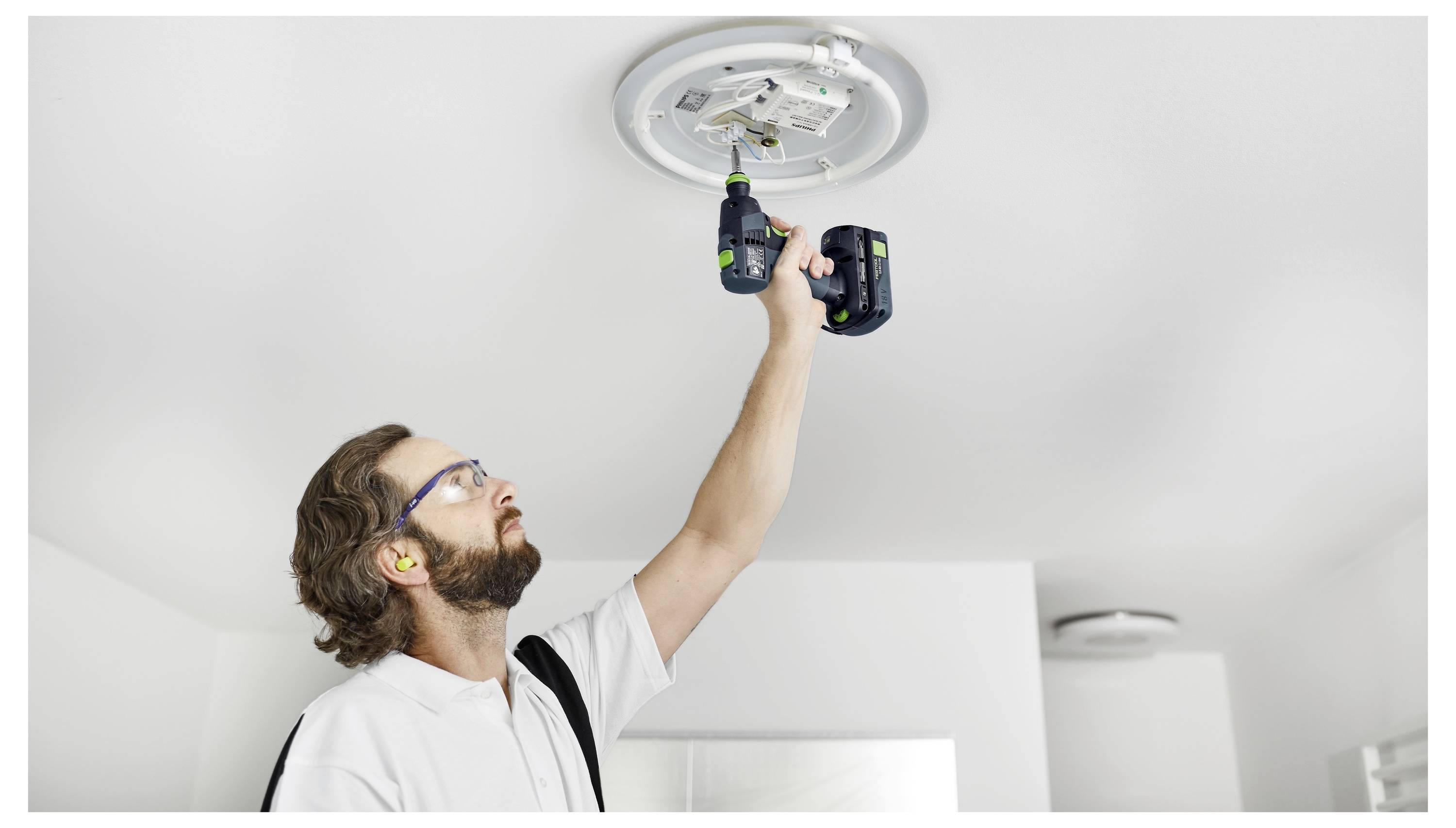 Eine Person verwendet einen Elektroschrauber, um ein Deckengerät zu installieren oder zu reparieren, wobei sie eine Schutzbrille und Ohrstöpsel aus Sicherheitsgründen trägt.