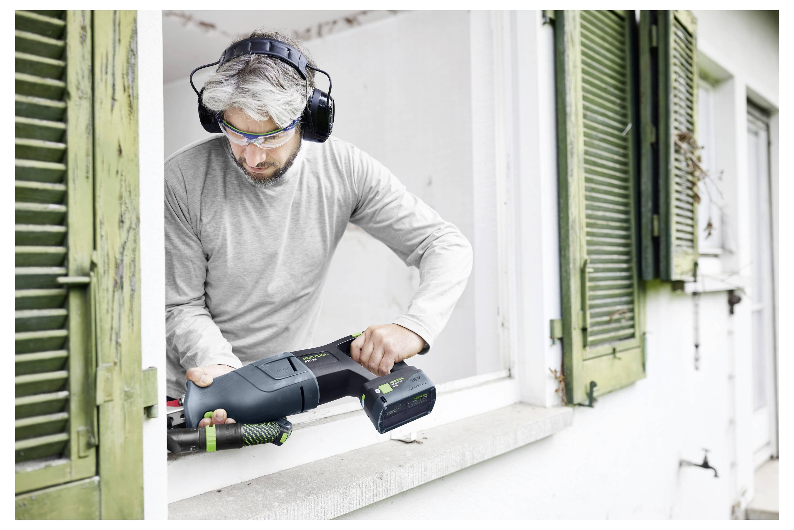 Eine Person in Schutzausrüstung nutzt ein Elektrowerkzeug, um durch die Fensterbank eines Hauses zu schneiden. Grüne Fensterläden sind an den Fenstern geöffnet.