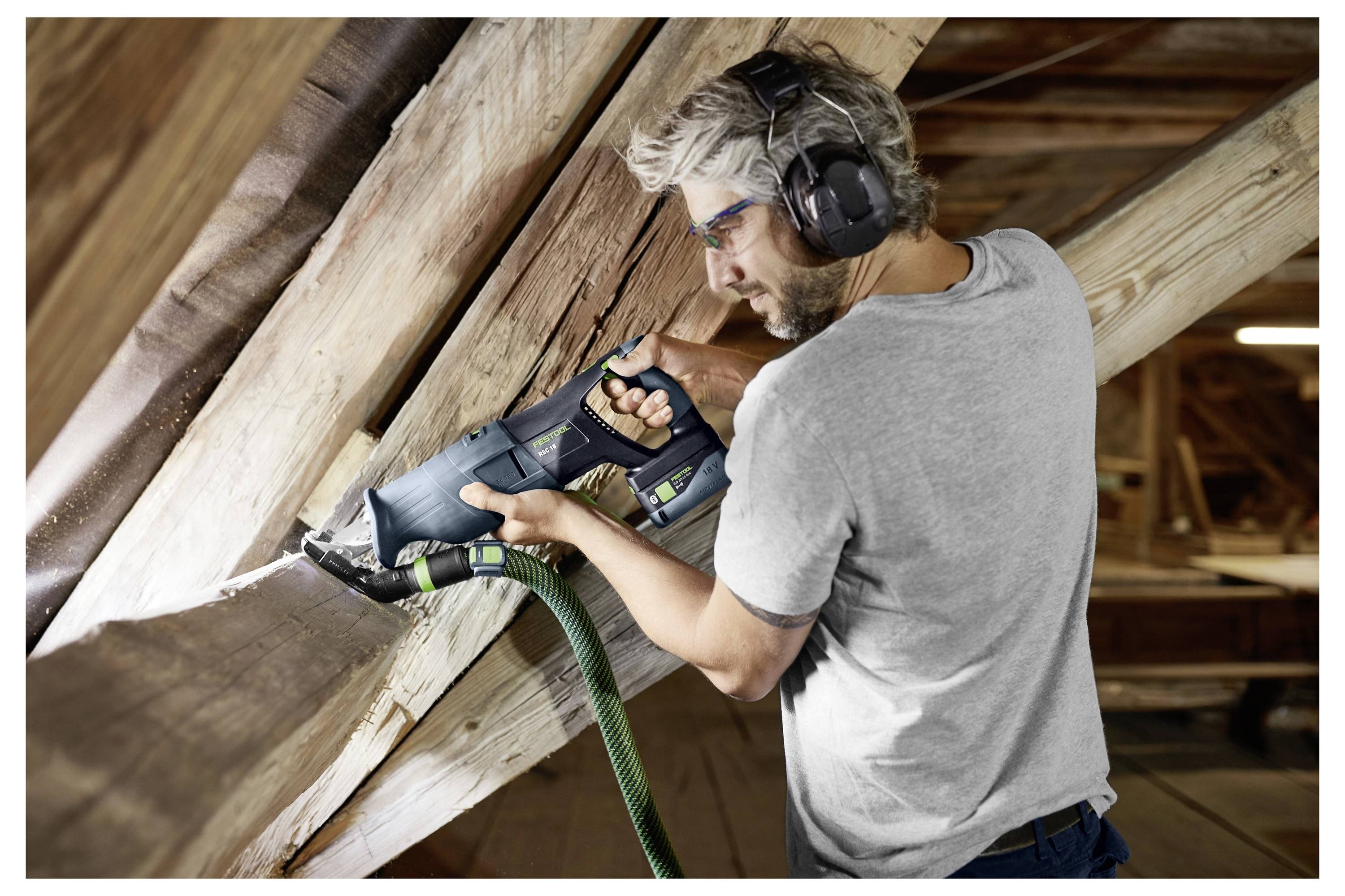 Eine Person in Schutzausrüstung nutzt eine Säbelsäge, um Holzbalken auf einem Dachboden oder in einem Baubereich zu schneiden.