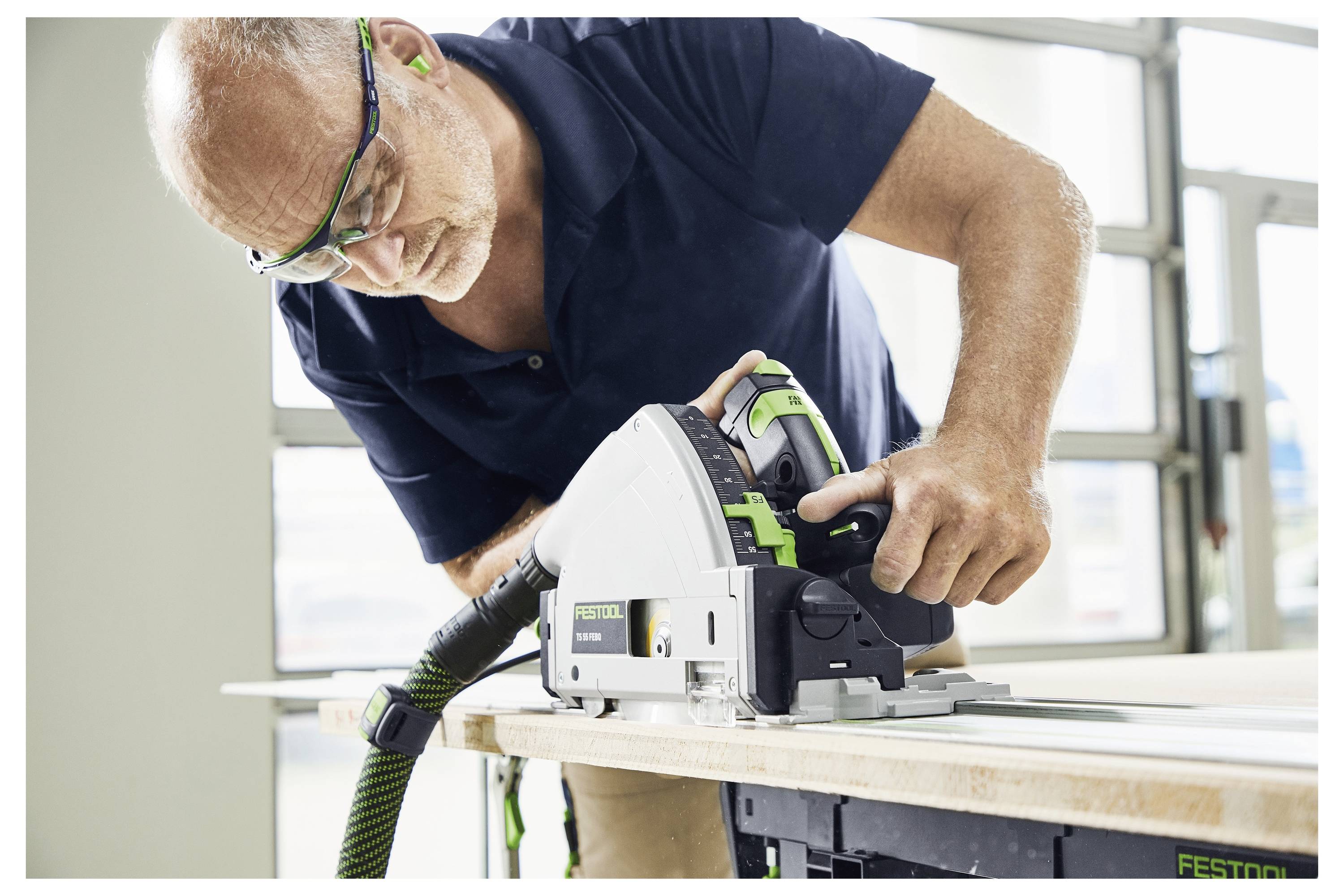 Ein Mann, der eine Schutzbrille trägt, verwendet eine Tauchkreissäge, um ein Stück Holz auf einem Tisch zu schneiden, und demonstriert dabei präzises Holzbearbeiten.