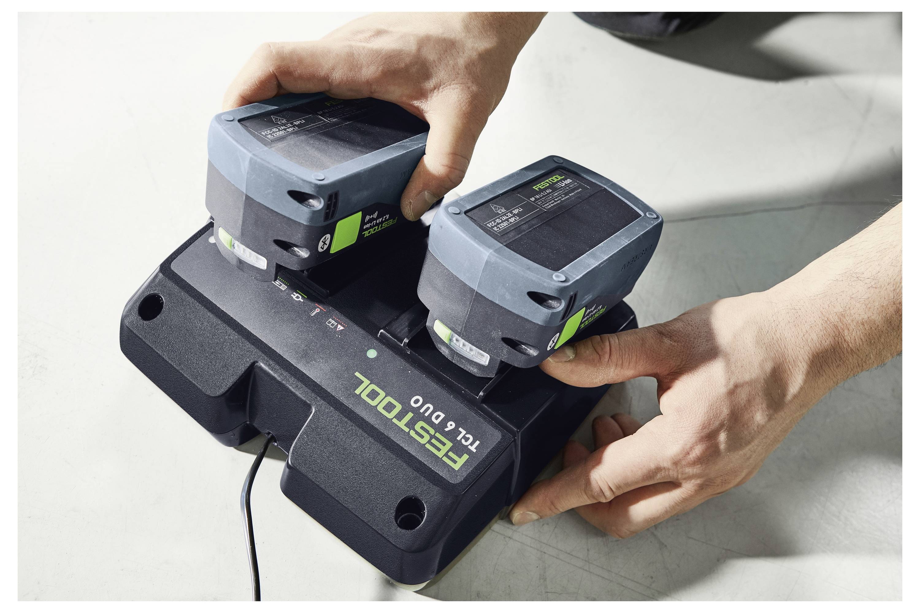 Zwei Hände platzieren zwei Batterien auf einem Festool TCL 6 Duo Batterieladegerät auf einer hellhellen Oberfläche.