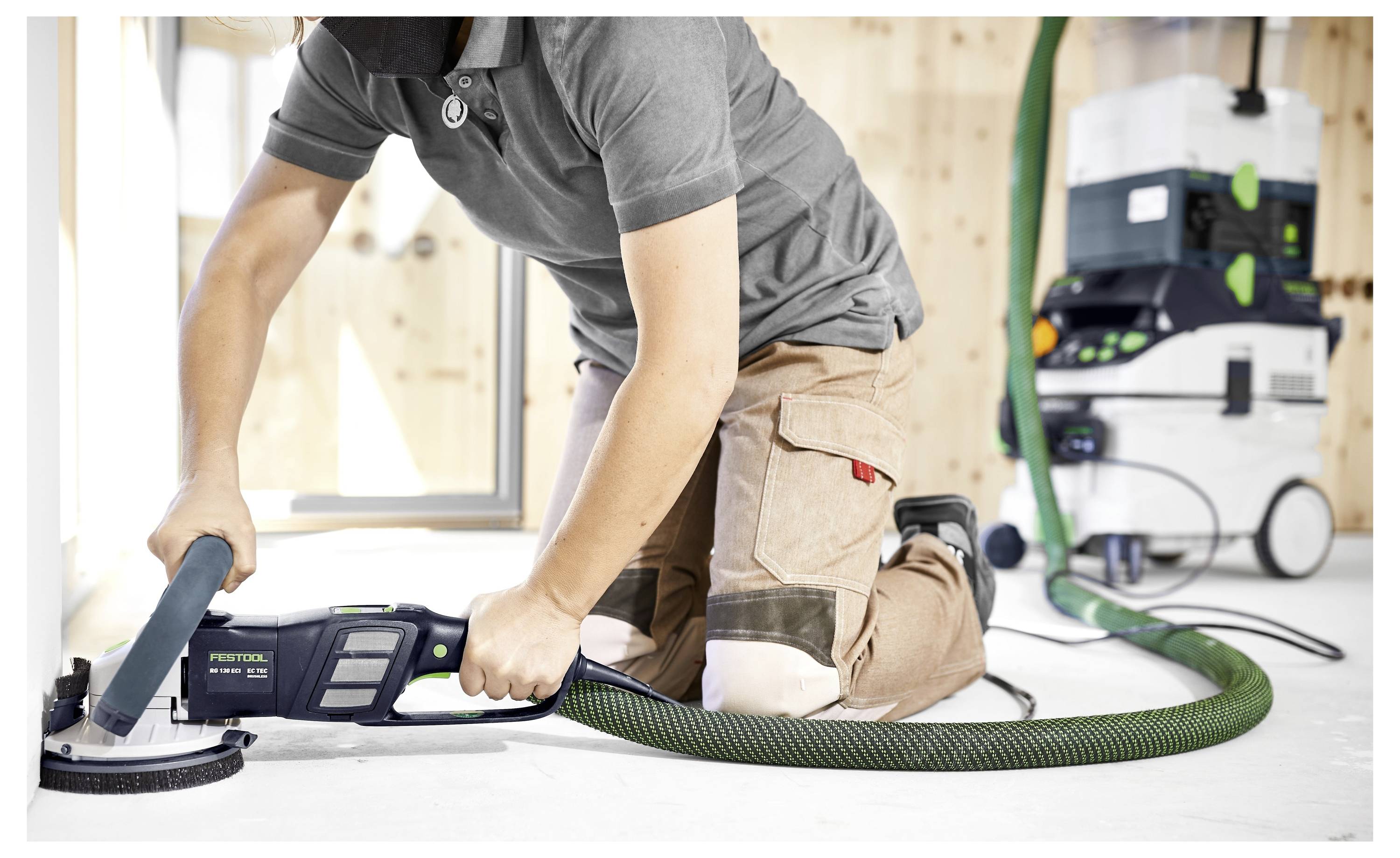 Eine Person kniet auf dem Boden und benutzt ein Elektroschleifsystem mit Absauganlage auf einer Holzoberfläche, wobei ein Staubabsauggerät durch einen grünen Schlauch angeschlossen ist.