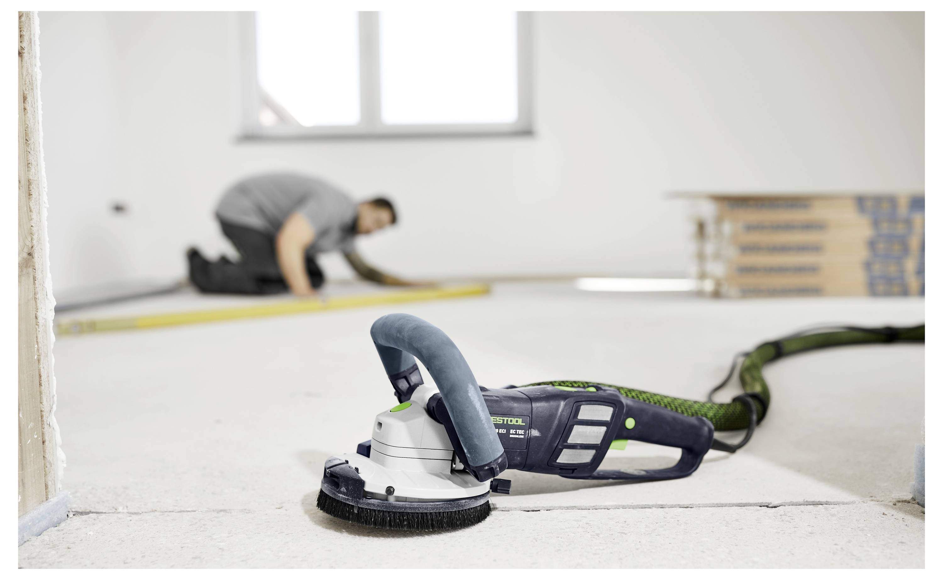Eine Person kniet nieder und misst einen Raum mit einem Stahllineal im Hintergrund; ein Elektroschleiffer ruht im Vordergrund auf dem Boden.