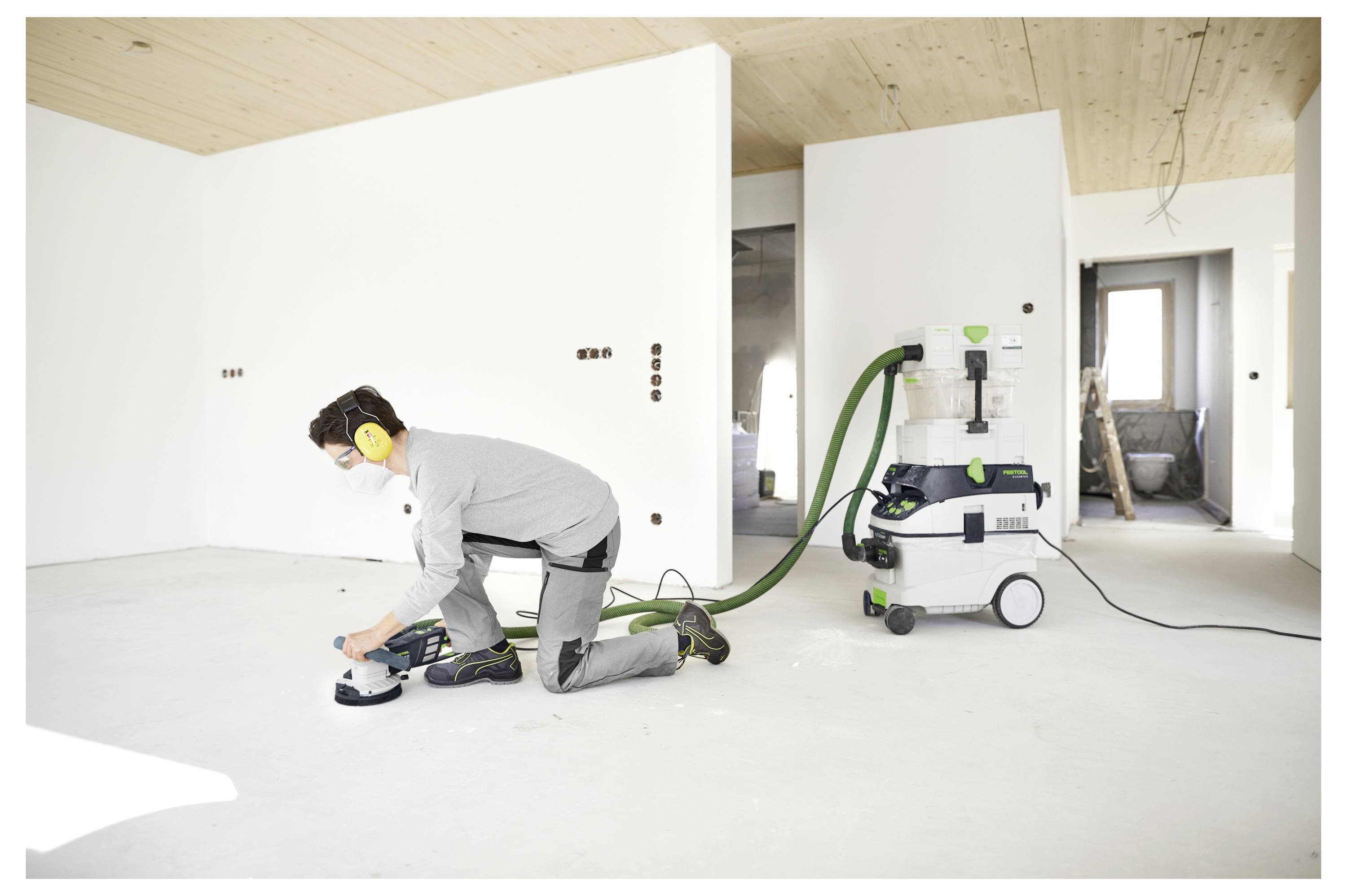 Arbeiter in Schutzausrüstung, der einen Boden mit einem angeschlossenen Staubextraktionssystem in einem teilweise fertiggestellten Raum mit weißen Wänden schleifen.