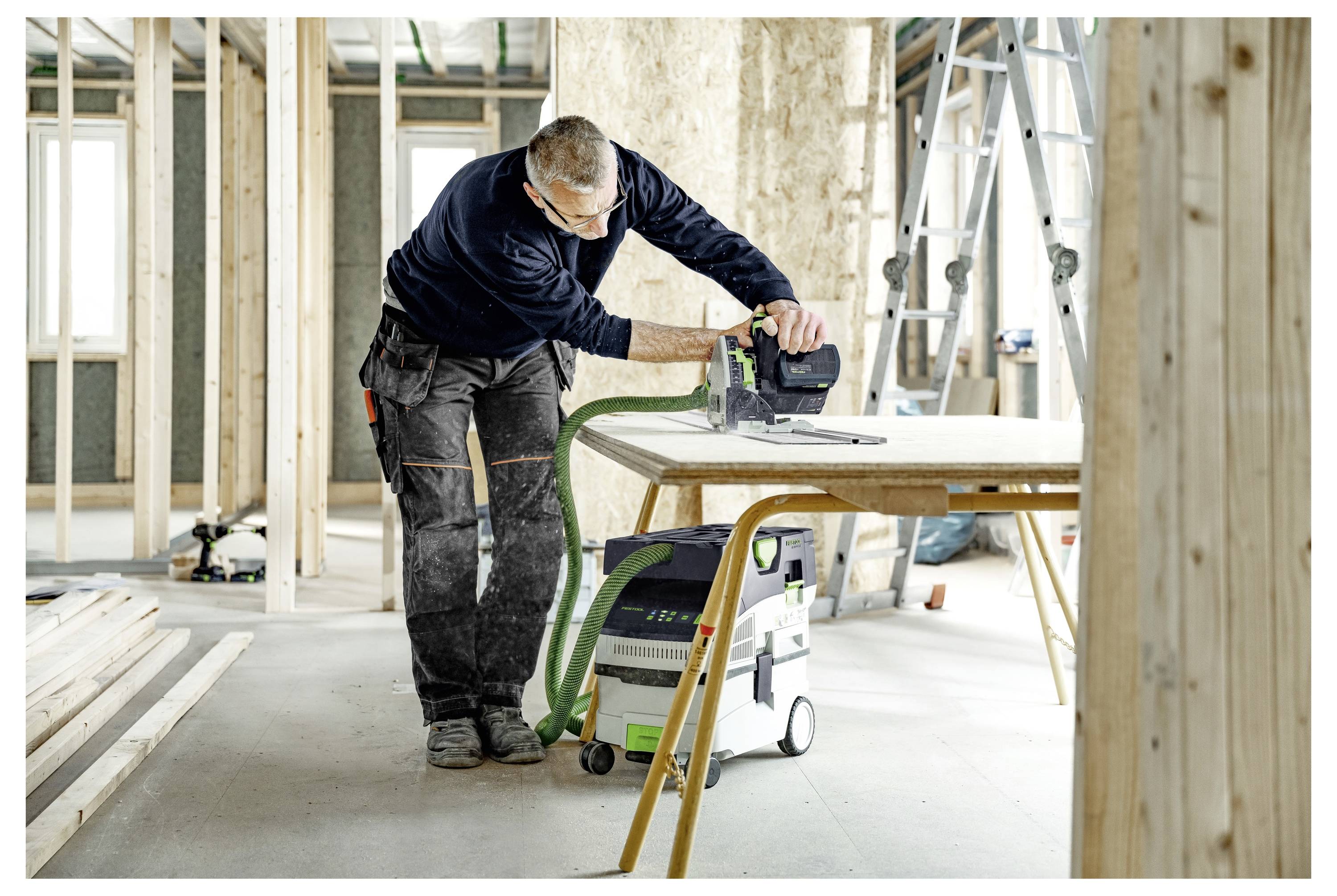 Eine Person benutzt ein Elektrowerkzeug auf einer Holzplatte auf einer Baustelle mit freiliegenden Holzbalken, einer Leiter und Baumaterialien in der Umgebung.